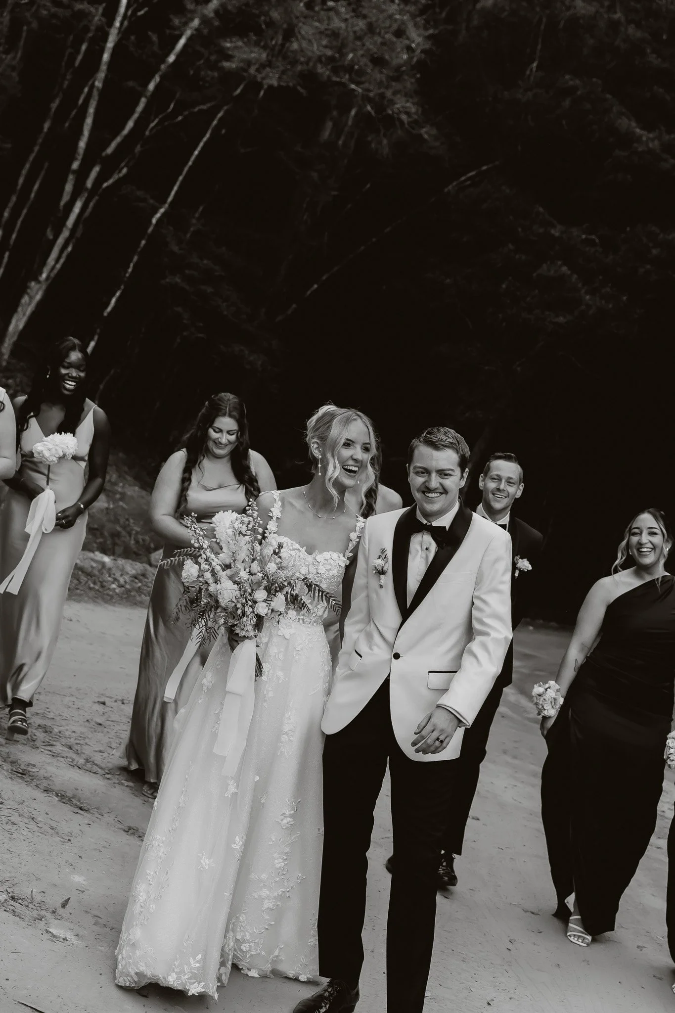 Black and white photo of a wedding party walking outdoors, with a bride in a lace gown holding a bouquet and a groom in a tuxedo, smiling. Bridesmaids and groomsmen walk alongside, smiling and laughing, in a natural setting with trees in the background.