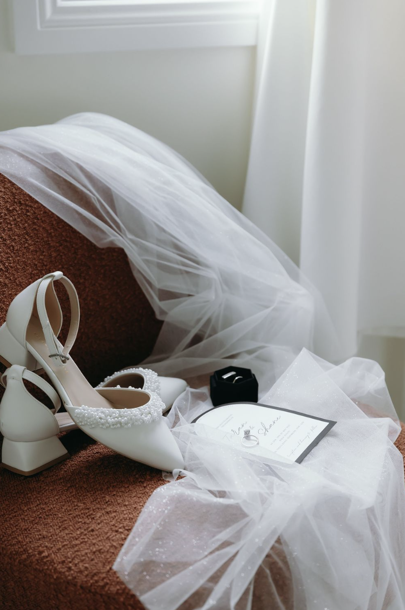 Wedding shoes, a wedding ring, a thank you card, and a small black box arranged on a brown cloth surface, with white tulle fabric draped nearby.