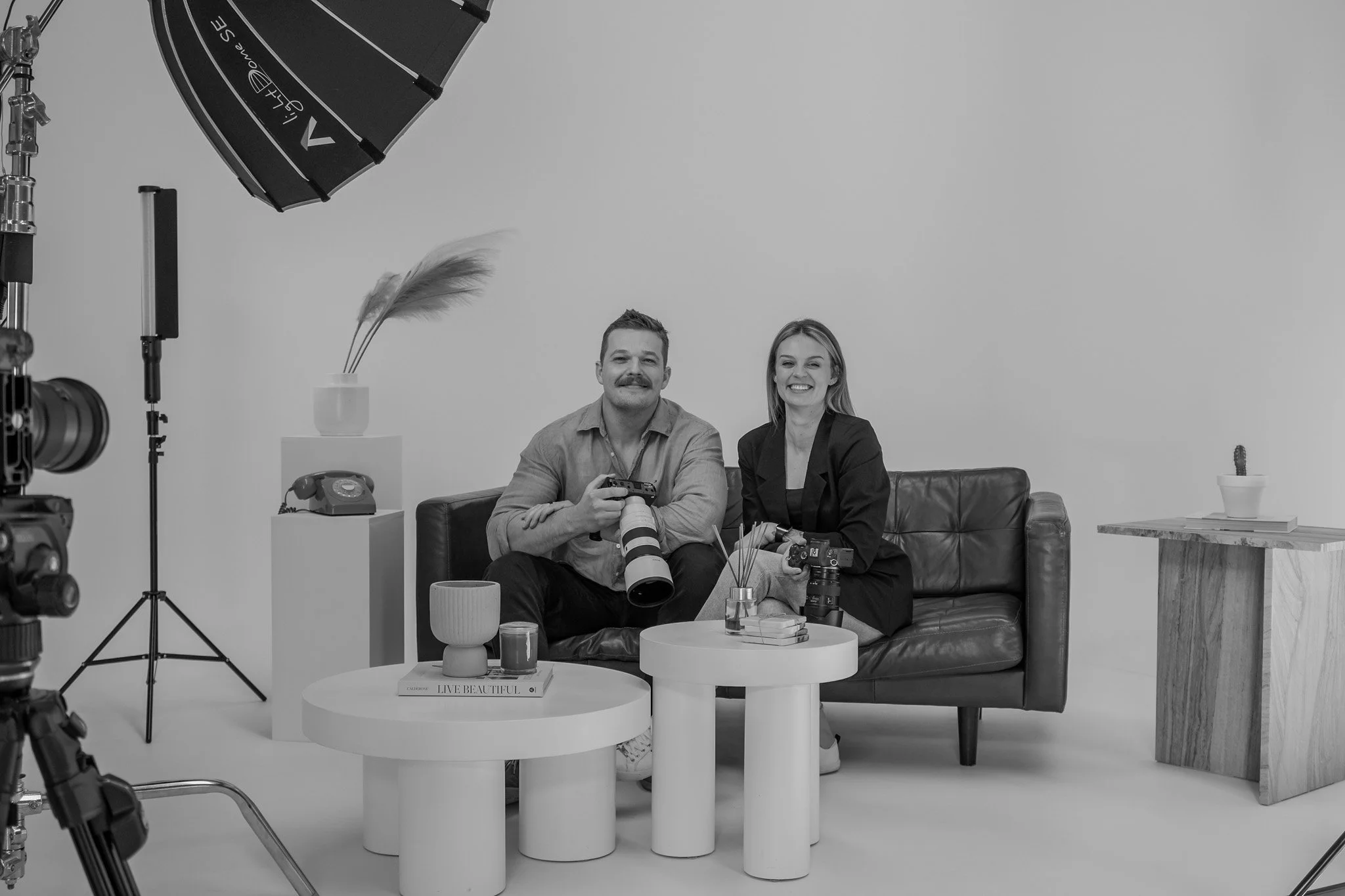 Two people sitting on a black leather couch in a photography studio, smiling at the camera. The man on the left holds a professional camera with a large lens, and the woman on the right holds a camera in her lap. Studio equipment and decor, including a large umbrella light, a small side table with a cactus, and a few decorative books, are visible in the background.