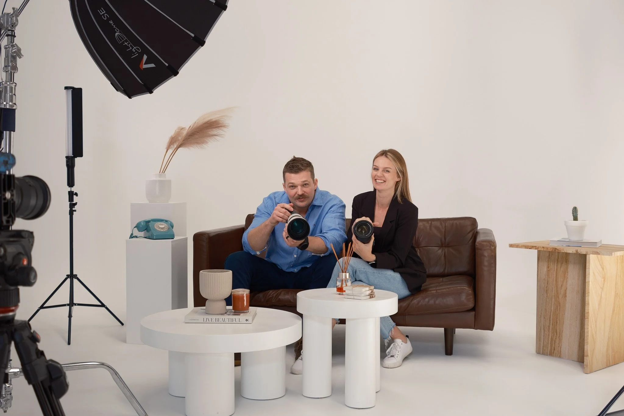 Two people, a man and a woman, sitting on a brown sofa in a photography studio, holding cameras, smiling, with filming equipment around them, including a large black umbrella light and a camera on the left.