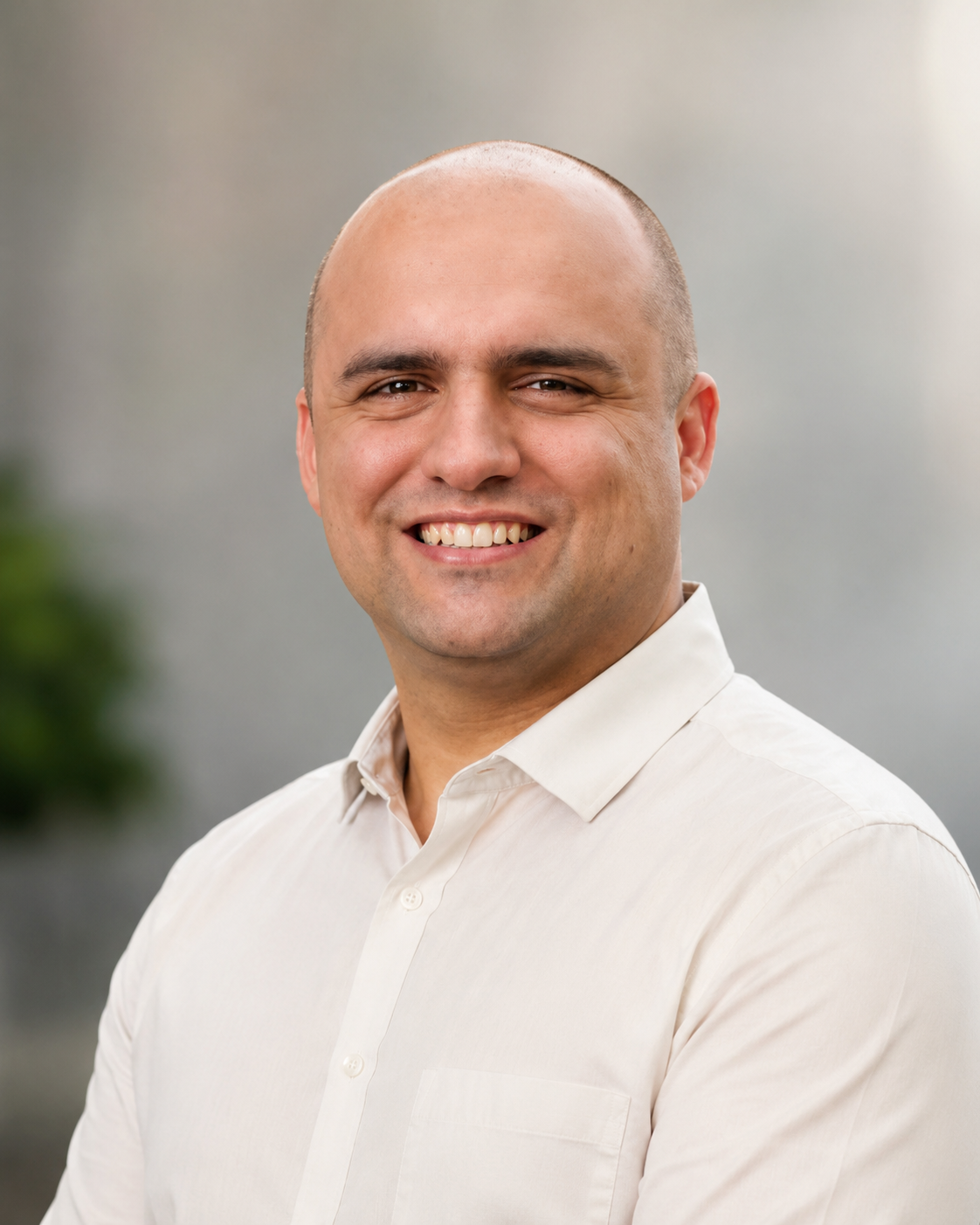 A smiling man with a shaved head wearing a light-colored dress shirt, standing in front of a blurred gray background.