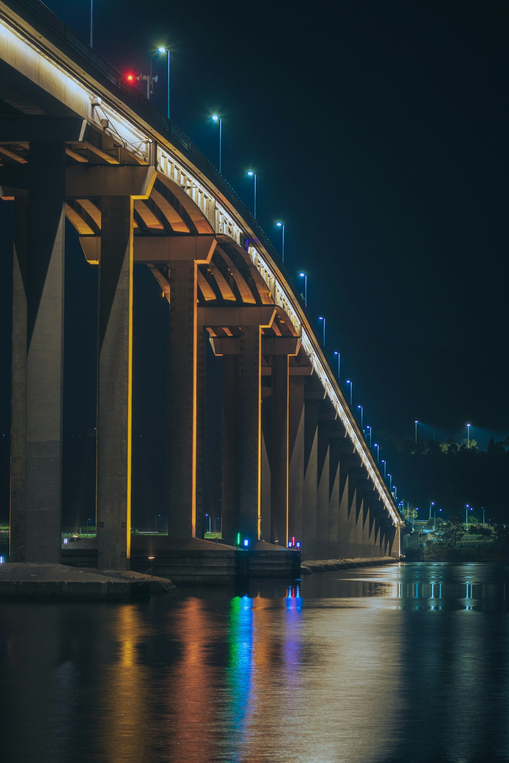 Night view of a bridge illuminated with blue lights, reflecting on the water below.