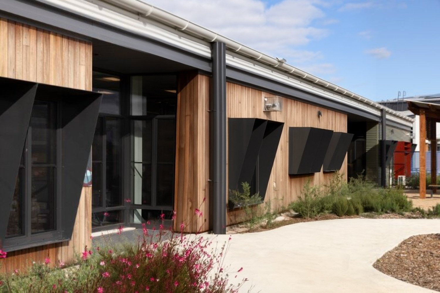 A modern building with wood siding, black window awnings, and drainage pipes, surrounded by landscaped plants and a concrete walkway under a partly cloudy sky.