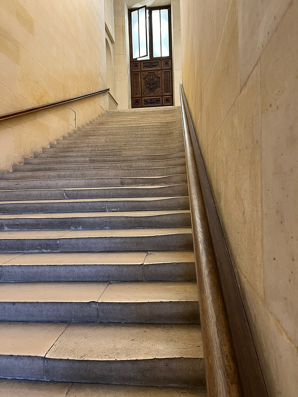 A staircase with stone steps and a wooden handrail leading up to an open door with intricate wood carvings, above which is an open window letting in natural light.