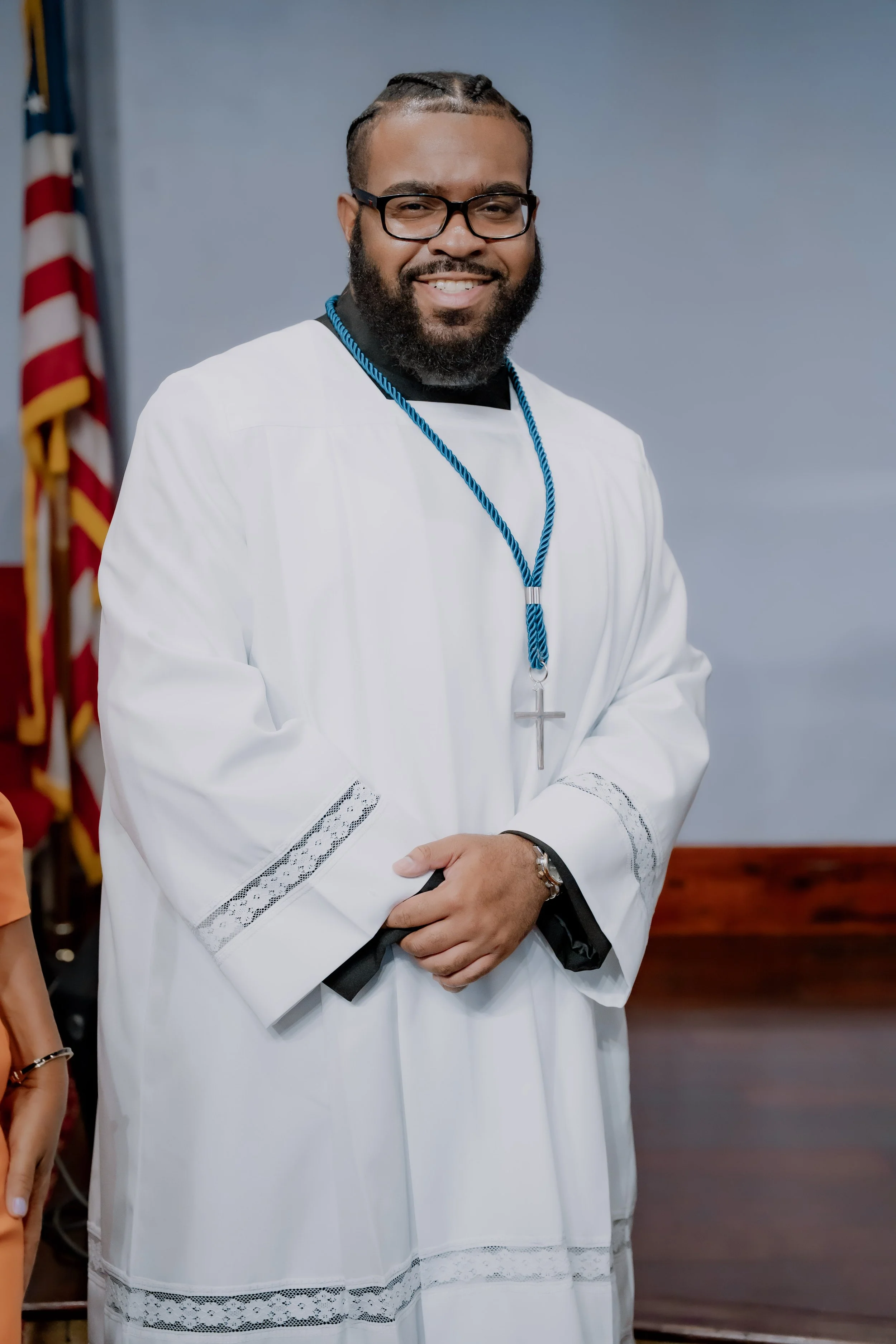A man wearing glasses and a white religious robe with lace details, standing indoors with an American flag in the background, smiling at the camera.
