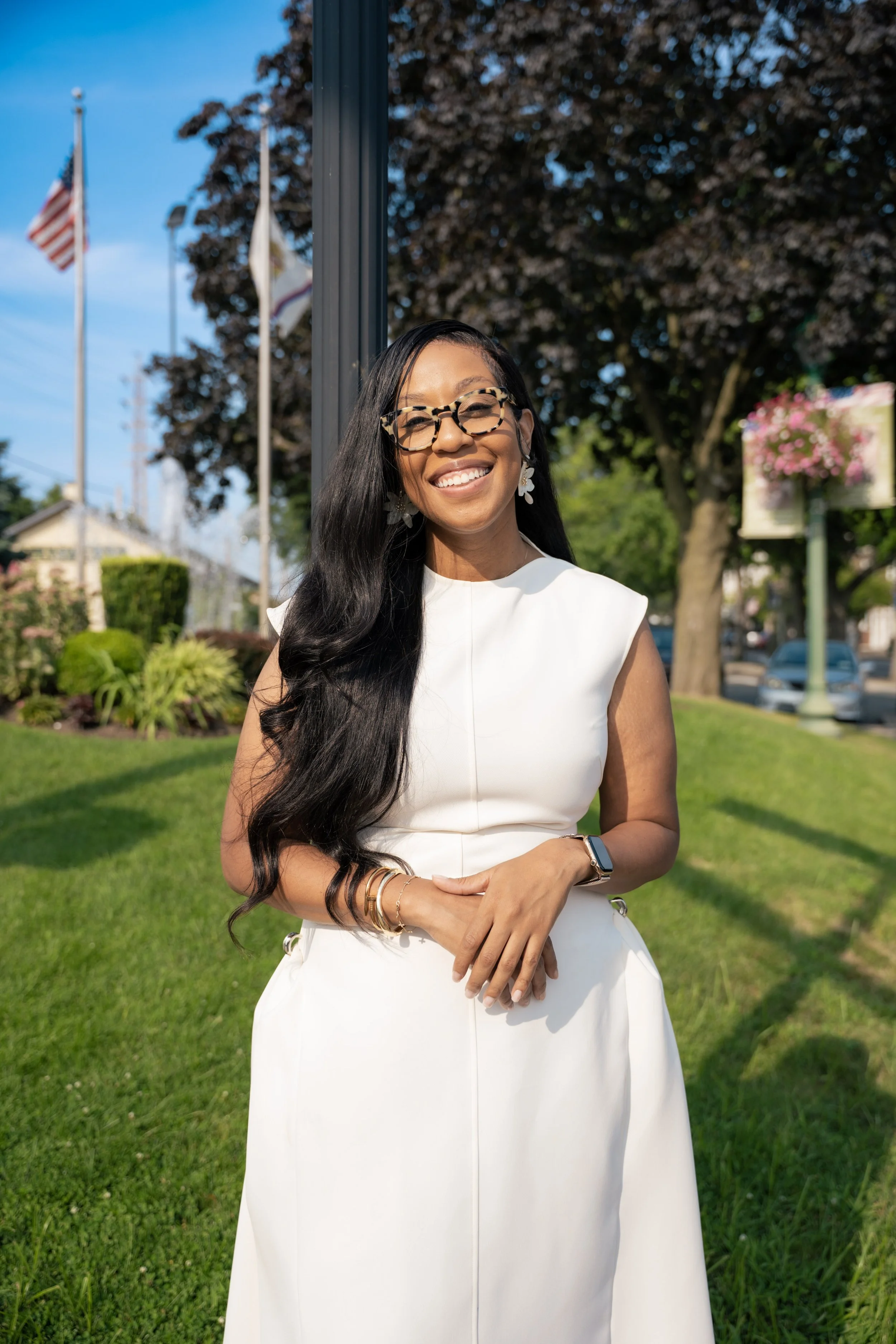 Smiling woman in white dress standing outdoors in a park, with trees, flowers, and American flag in the background.