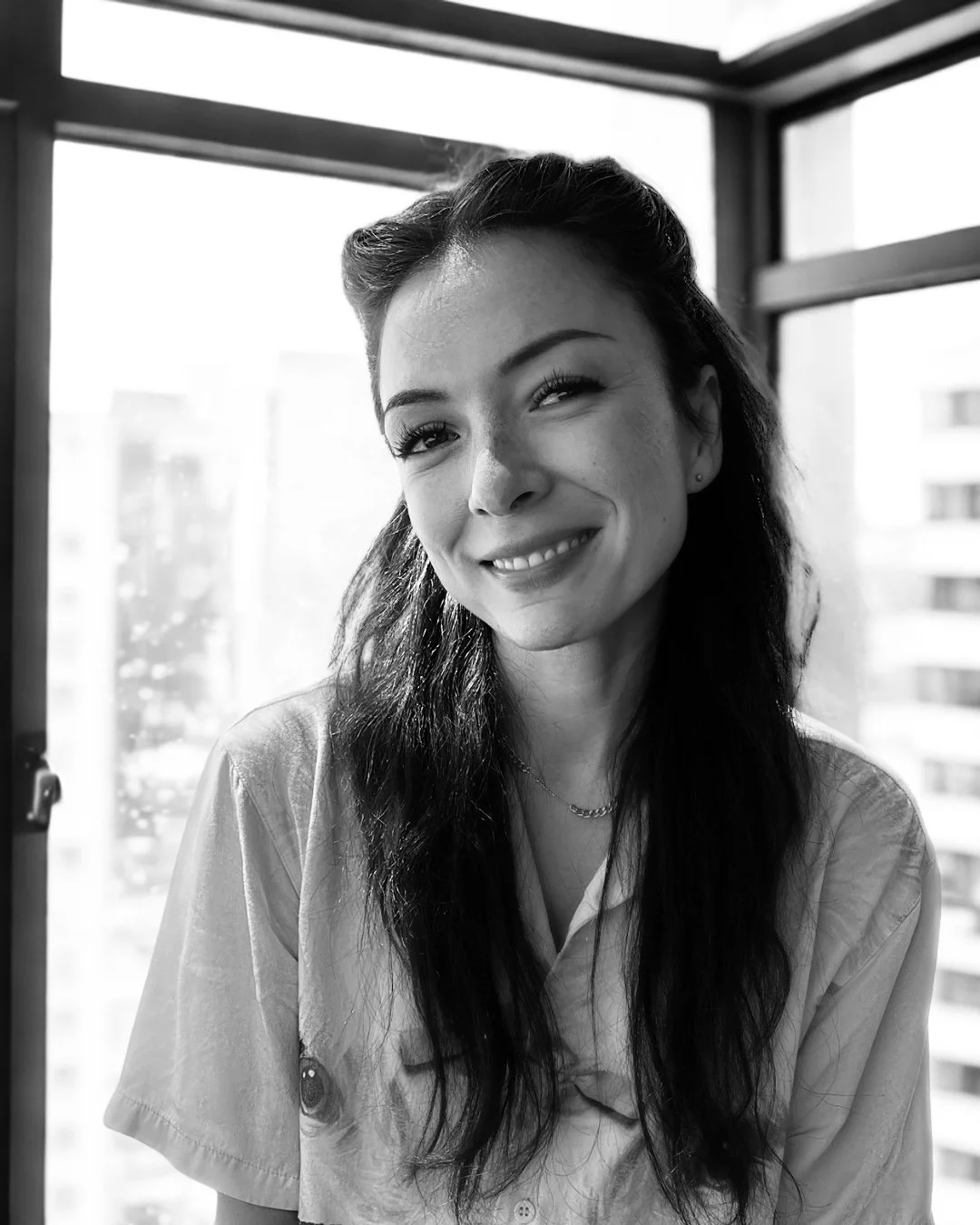 A woman with long dark hair, smiling and winking in front of a window, black and white photo.