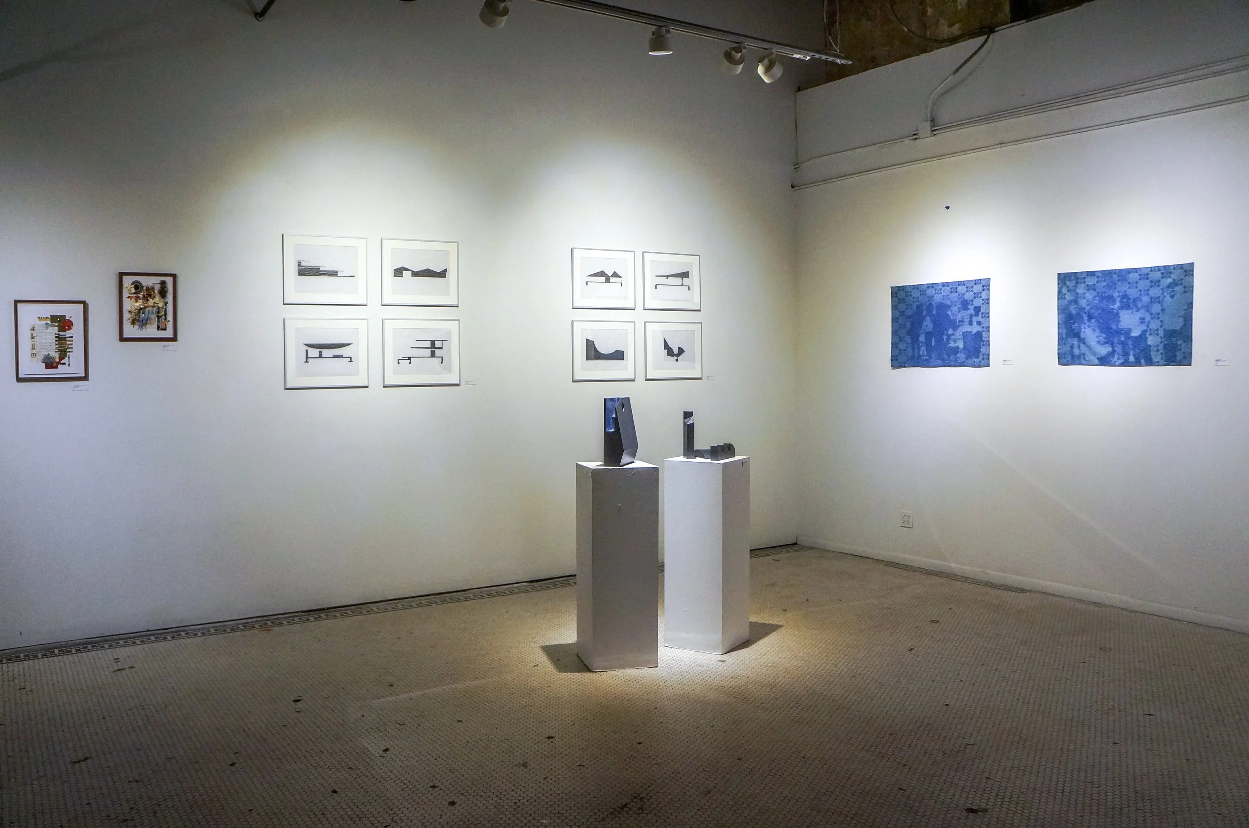 Art gallery with framed abstract art on white walls and blue patterned paintings, two sculptures on white pedestals, and overhead lighting.
