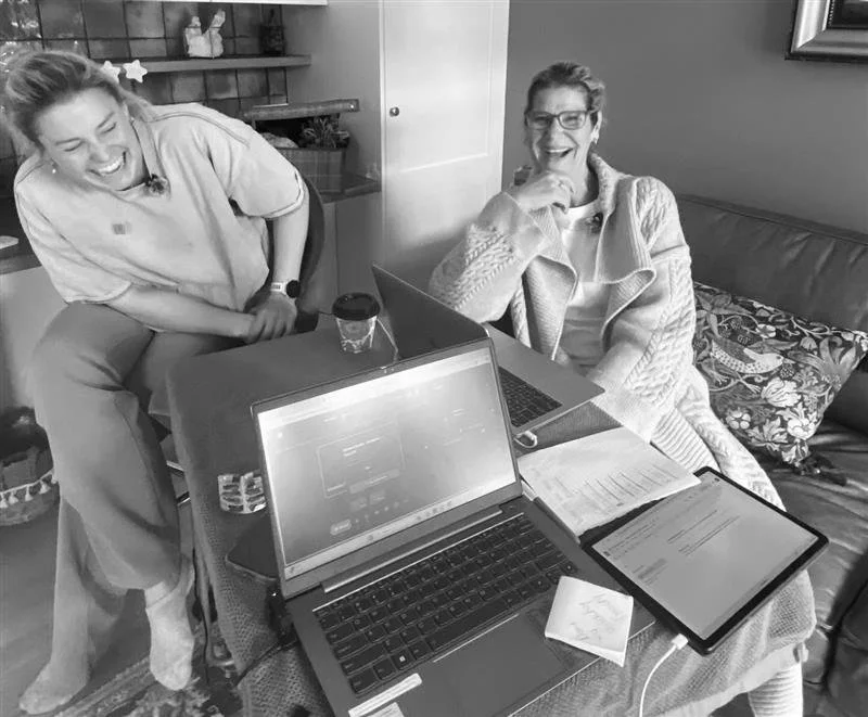 Two women are laughing and having a conversation in a cozy room. One woman is seated at a table with laptops, tablets, notebooks, and a cup of coffee, while the other woman is standing nearby, leaning on the table. The setting appears to be a casual meeting or work session.