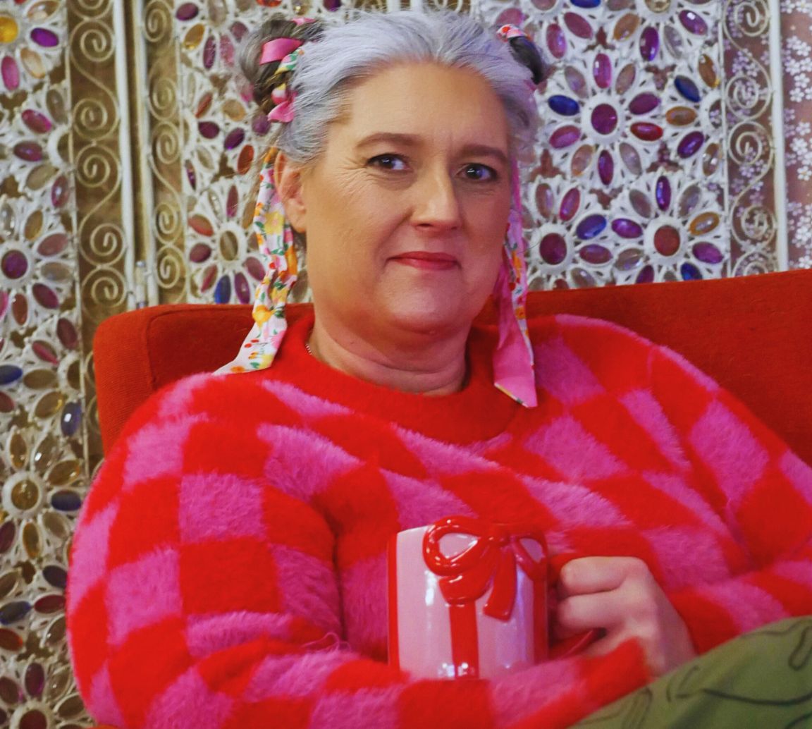 A woman with gray hair decorated with colorful ribbons, wearing a red and pink striped fuzzy sweater, sitting on a red chair with a decorative background. She is holding a small wrapped gift with a red ribbon.