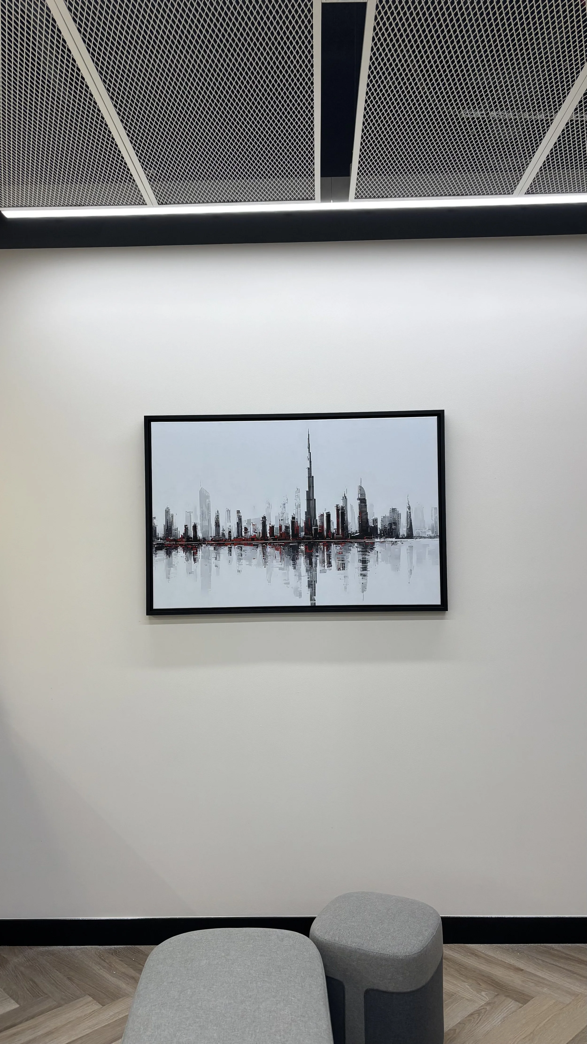 Minimalist interior office kitchen wall with a framed black and white Dubai cityscape photograph, ceiling with patterned panels, and gray seating in the foreground.