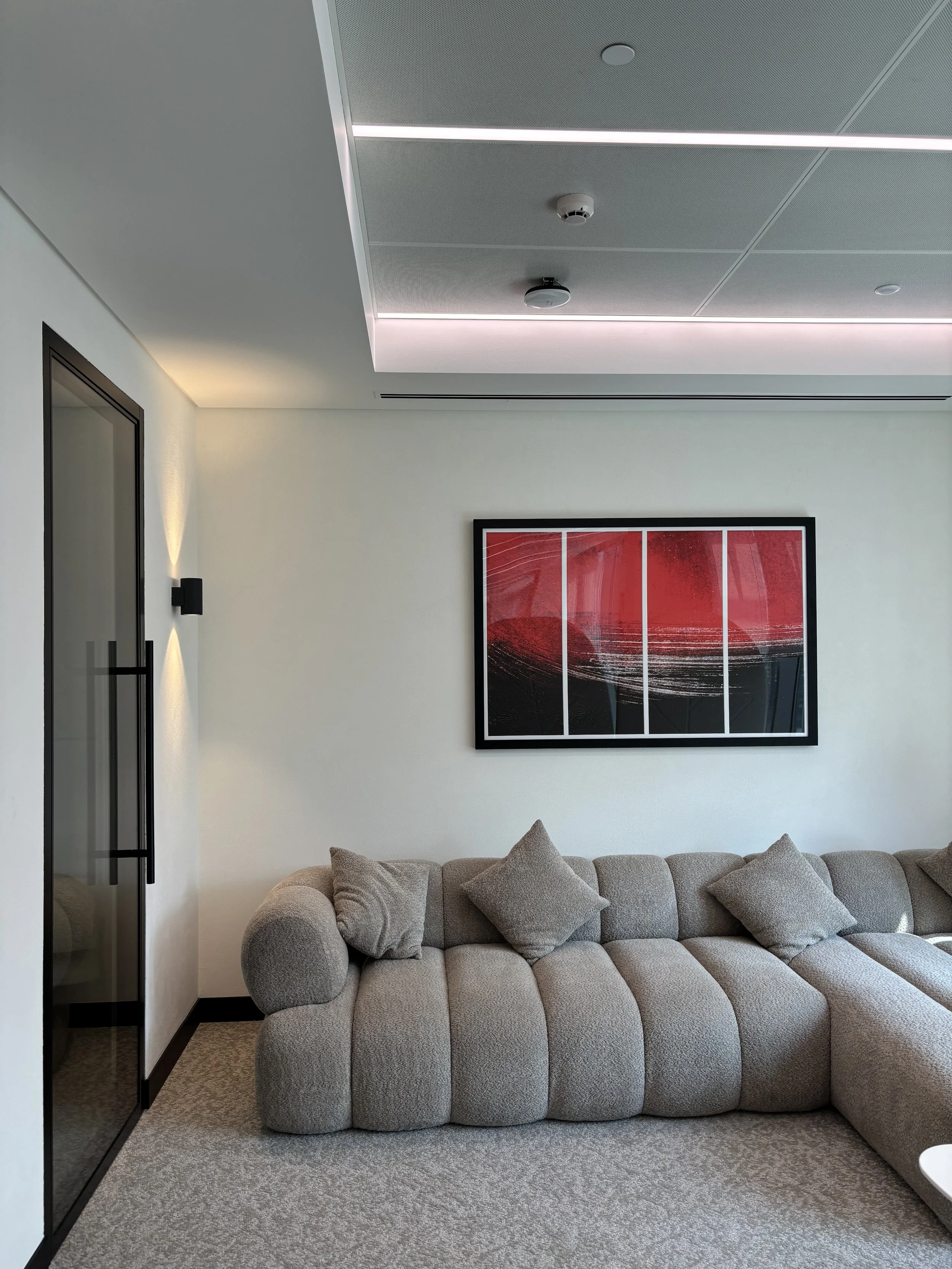 Modern office recreation room with beige sectional sofa and gray cushions, abstract red and black framed artwork on white wall, glass door, textured ceiling with subtle lighting, and neutral carpeted floor.