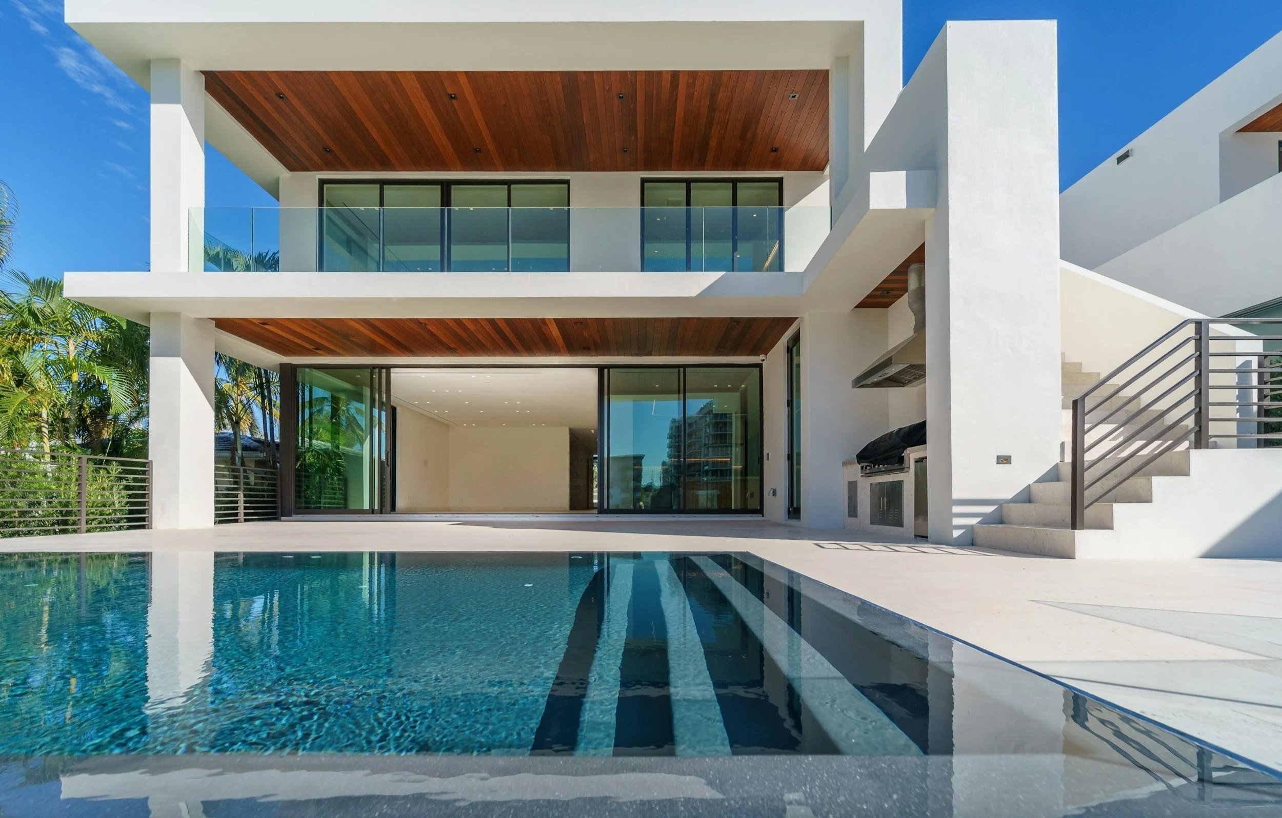 Modern two-story house with large glass sliding doors, a swimming pool in the backyard, and a balcony with glass railing on the second floor, surrounded by palm trees and clear blue sky.