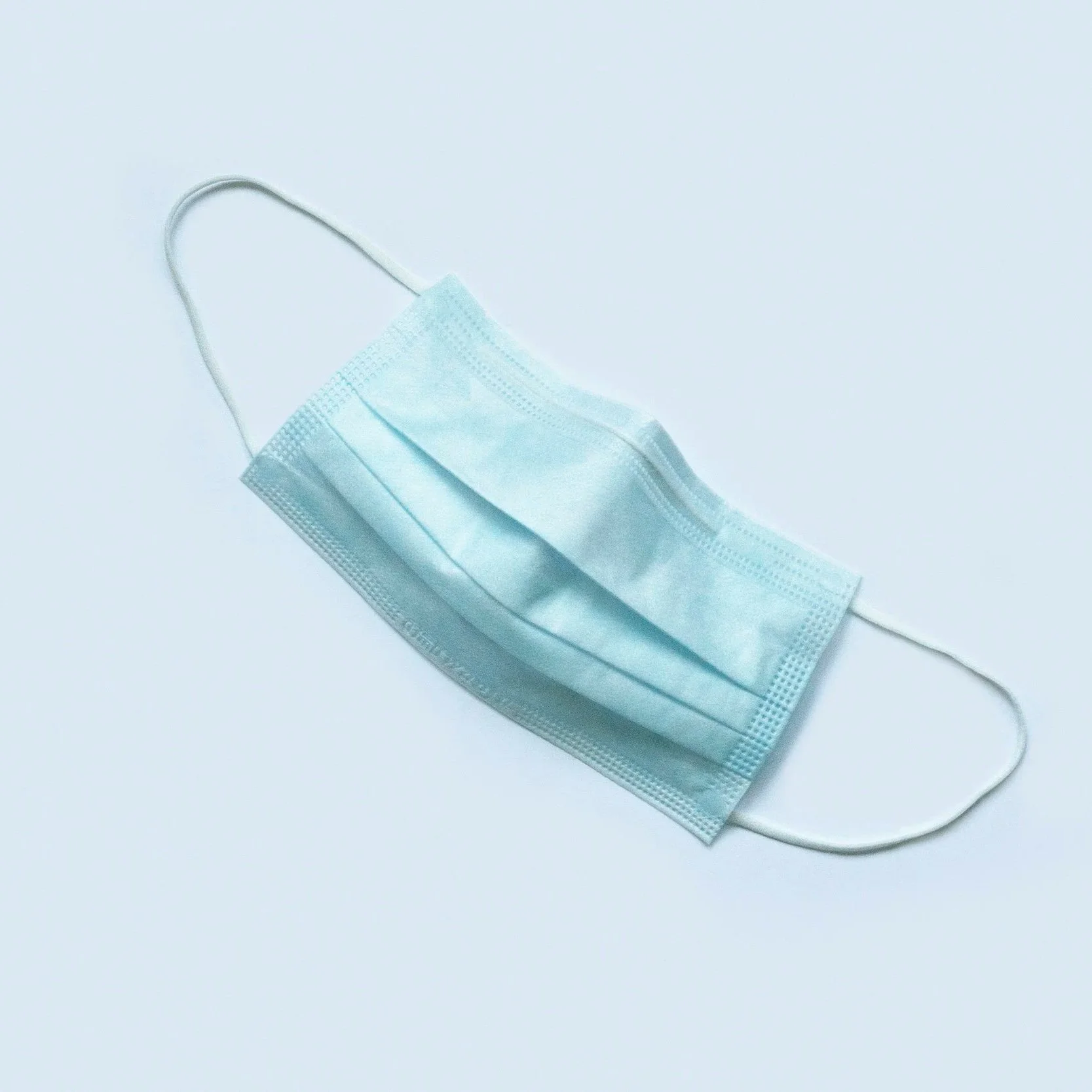 A light blue disposable face mask with ear straps on a plain white background.