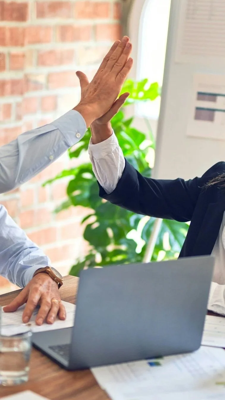 Two people giving each other a high five over a table with a laptop, documents, and a glass of water, in an office setting with a brick wall and green plant in the background.