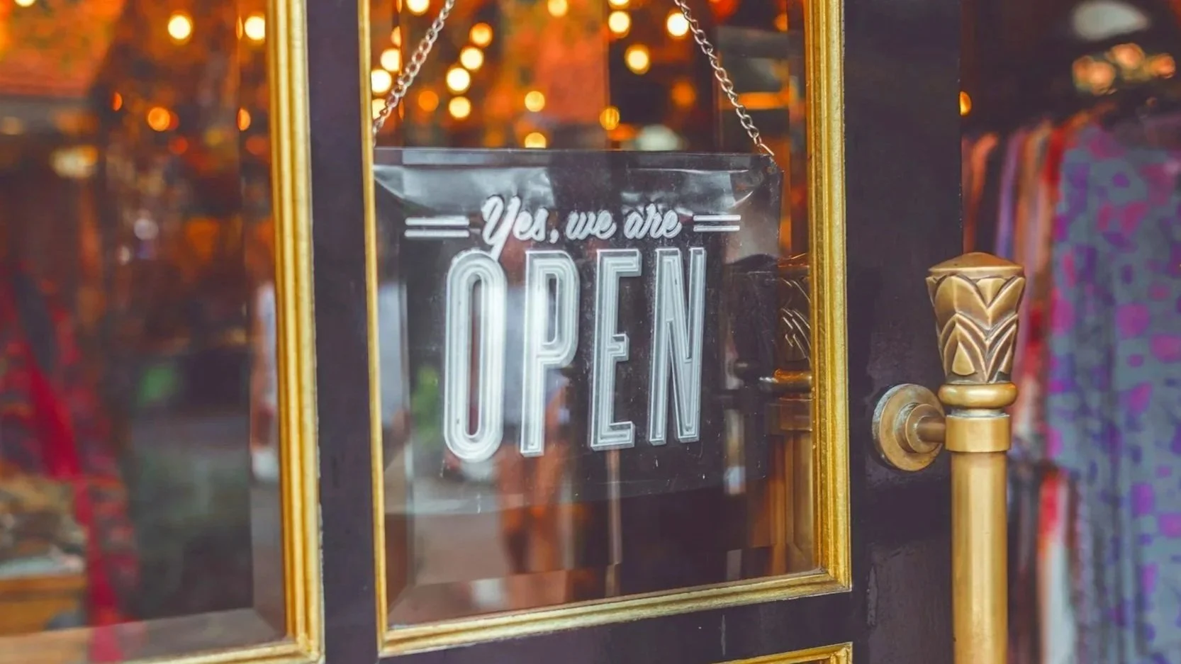 Glass door with an illuminated sign that reads, 'Yes, we are OPEN,' surrounded by gold trim, with indoor lights and clothing in the background.