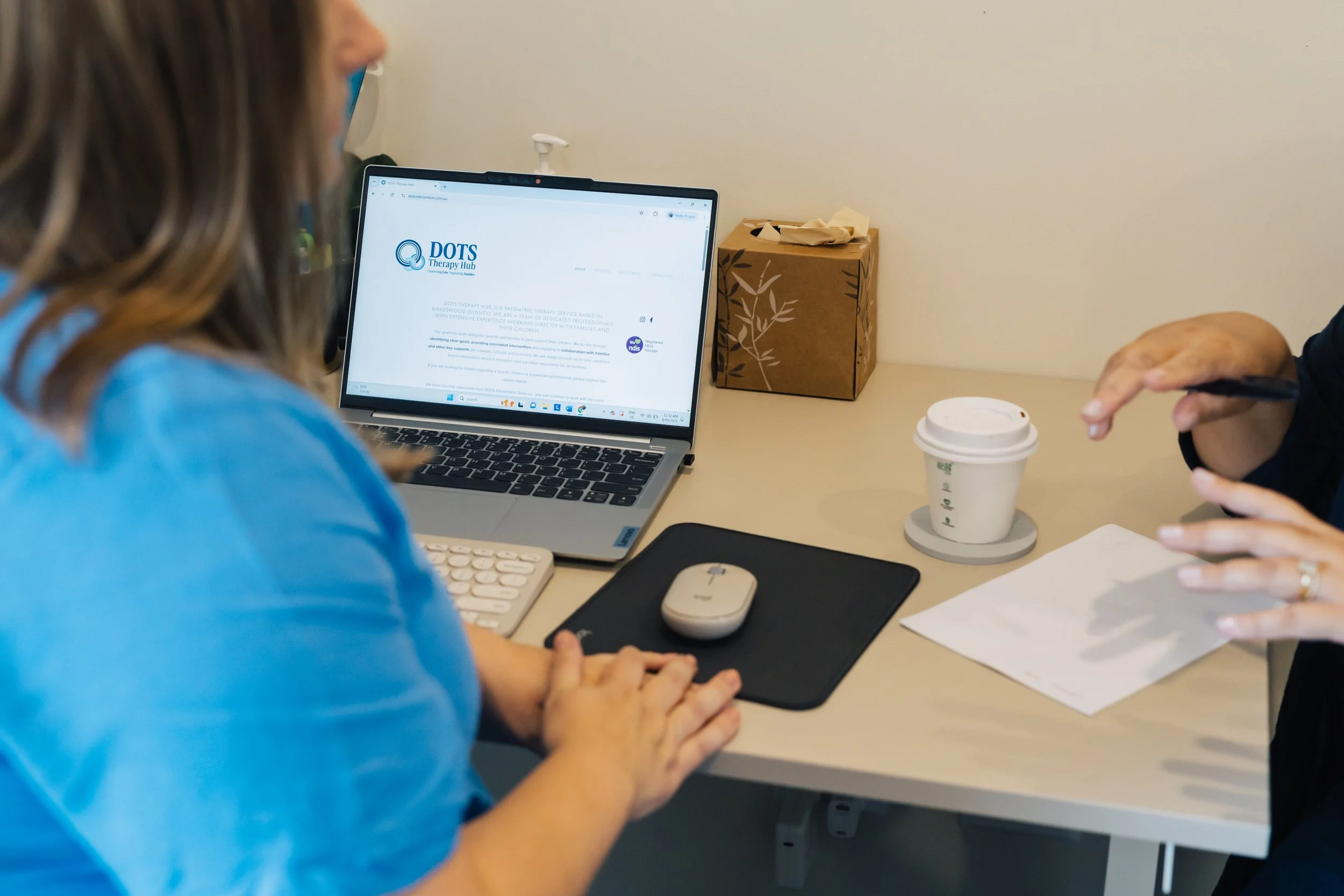 People sitting at a table in a meeting, one with a laptop displaying a website, and another with a coffee cup and papers.