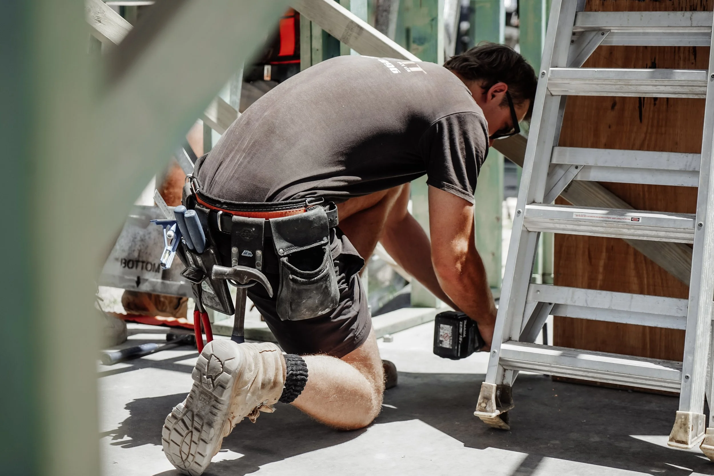 Builder kneeling on a residential renovation site in Hendra, Brisbane, using a power drill to secure building materials.