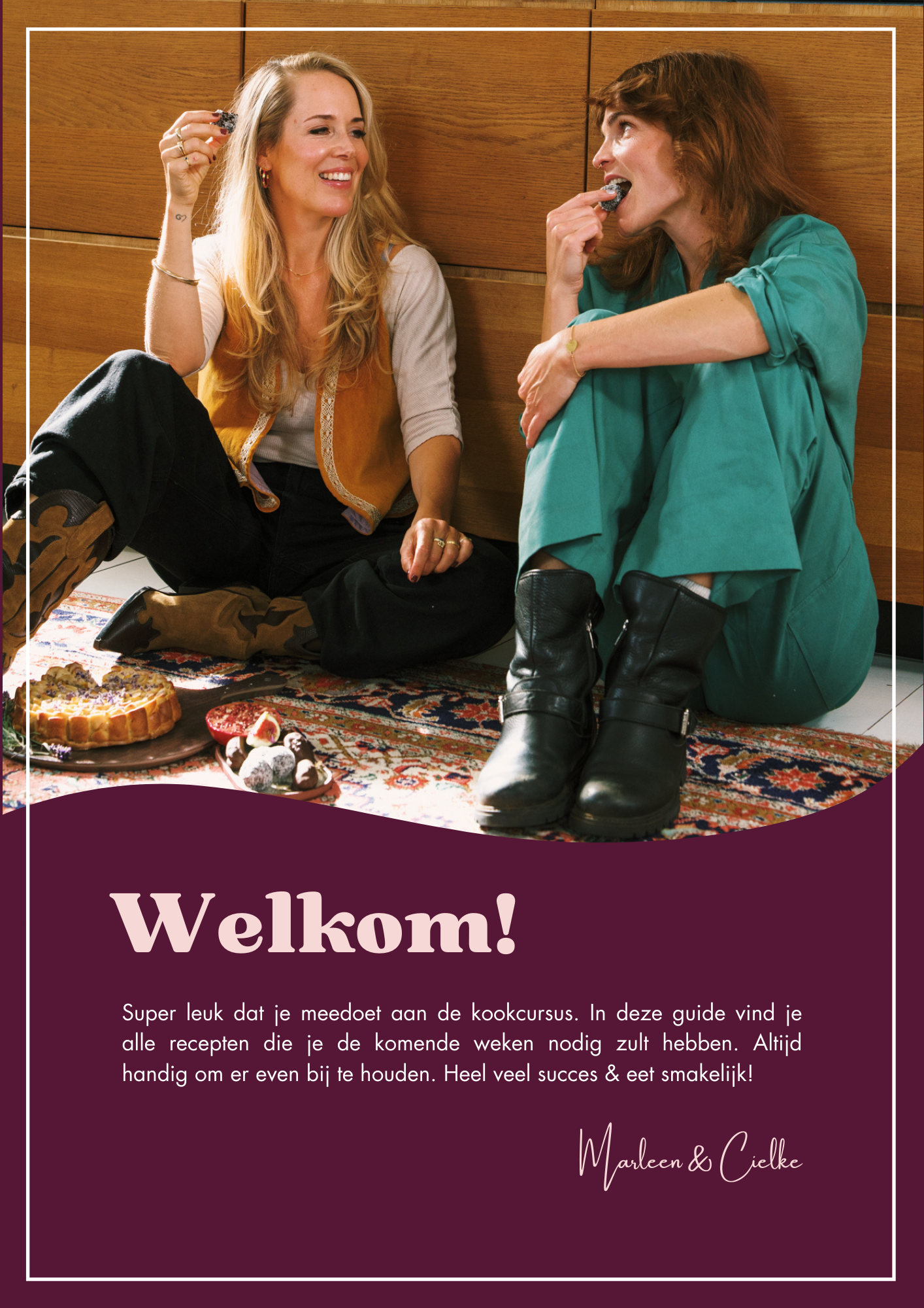 Two women sitting on the floor of a room with wooden wall panels, smiling and eating cookies, with cakes and sweets on the carpet in front of them. One woman has blonde hair and is wearing a beige vest over a white top, while the other has red hair a