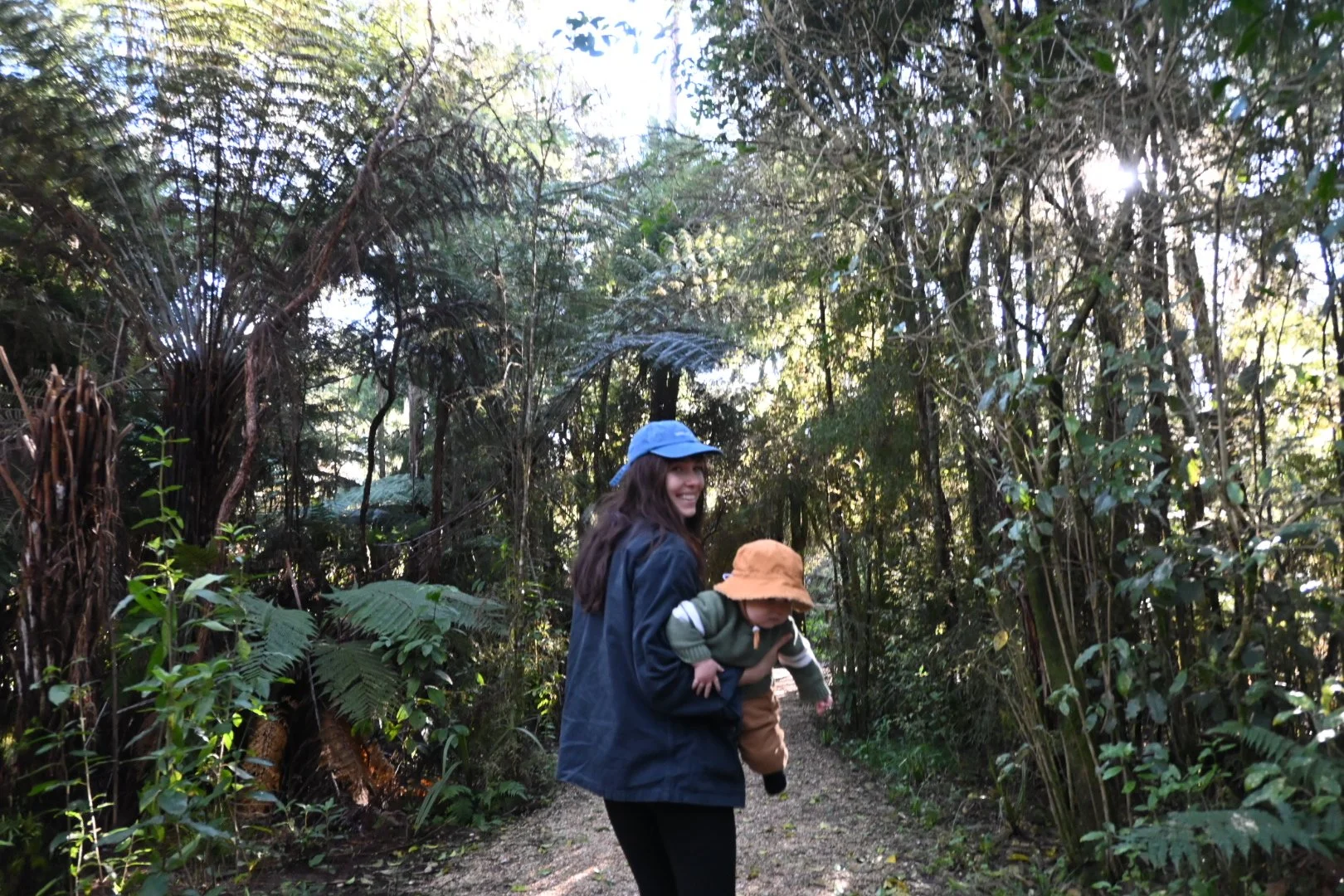 Smiling woman carrying a child in a forest trail surrounded by dense greenery and tall trees