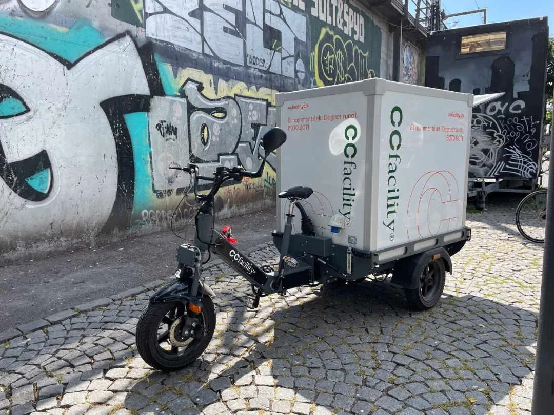 Cargo bike with storage box parked on cobblestone street, graffiti wall in background.