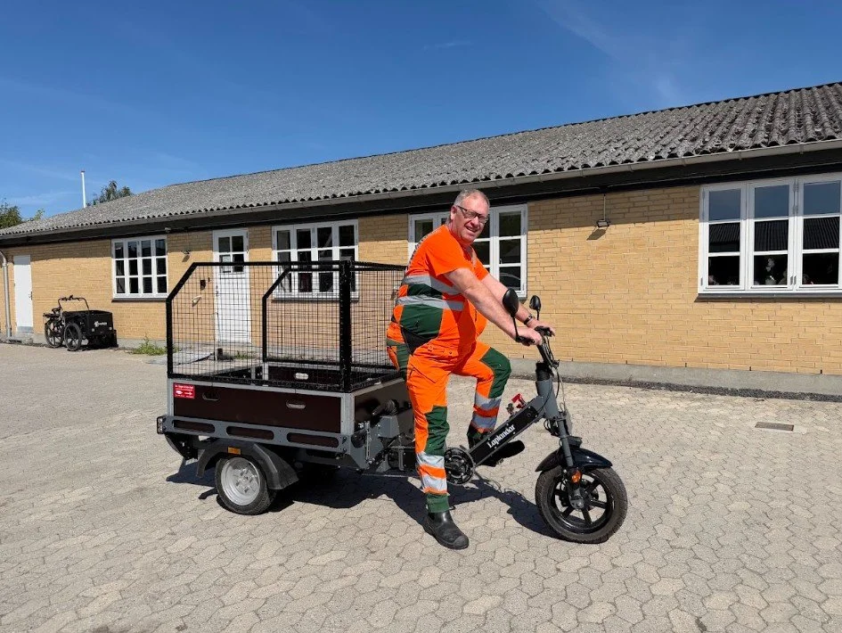 Kommunal medarbejder i arbejdstøj på Laplandar L-250 el-arbejdscykel med åben aluminiumskasse foran en driftsbygning.