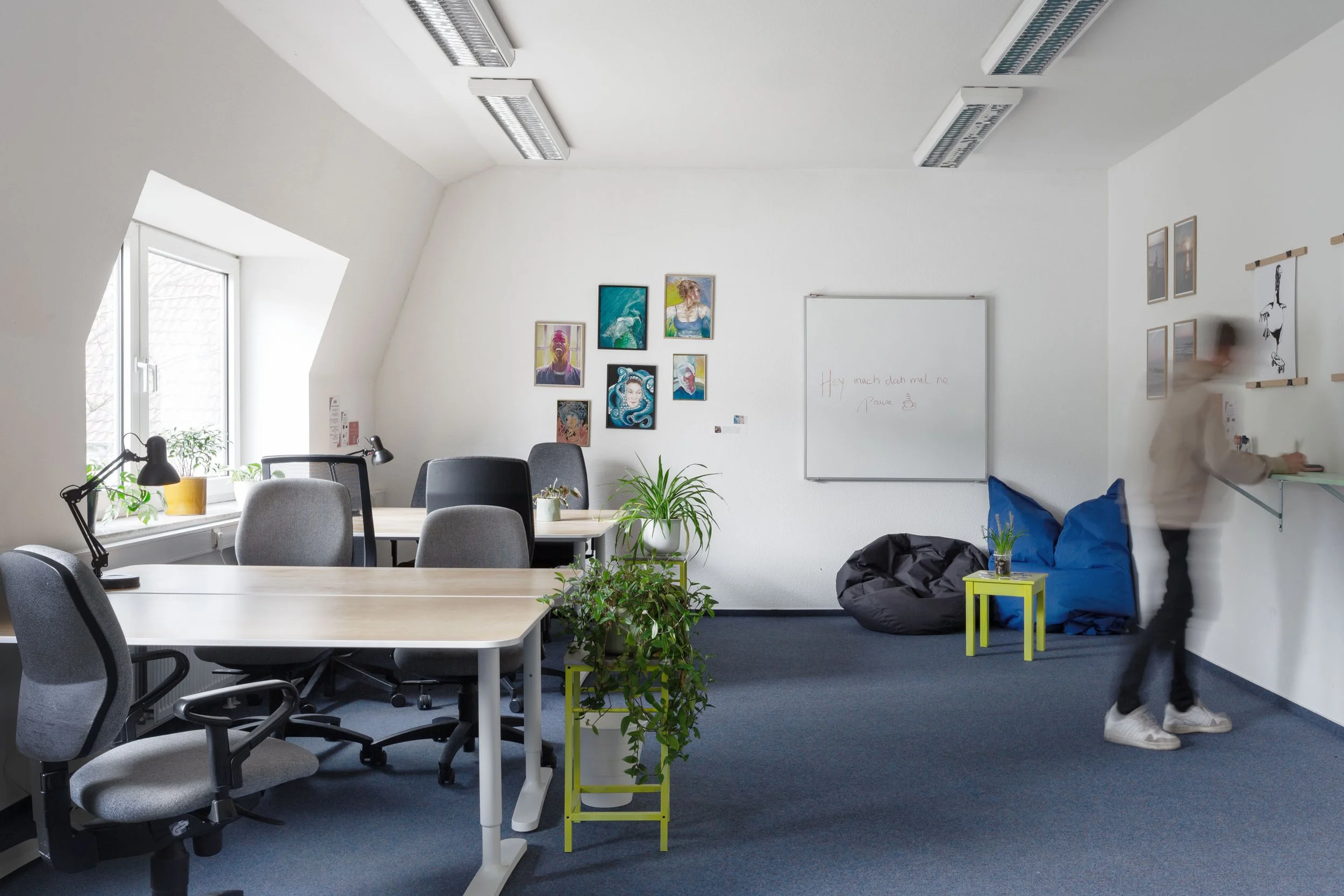 Auf dem Bild sieht man einen hellen großen Büroraum mit vielen Tischen und Stühlen. Rechts steht eine verschwommende Person.