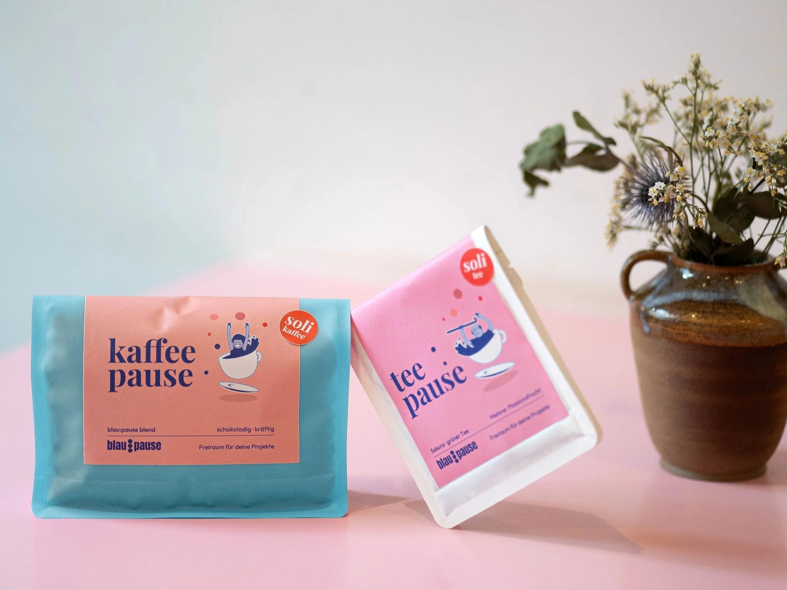 Auf dem Bild sieht man einen rosafarbenden Tisch. Auf dem Tisch steht ein Blumenstrauß in einer Tonvase. Daneben steht eine blaue Kaffeeverpackung und eine weiße Teeverpackung.