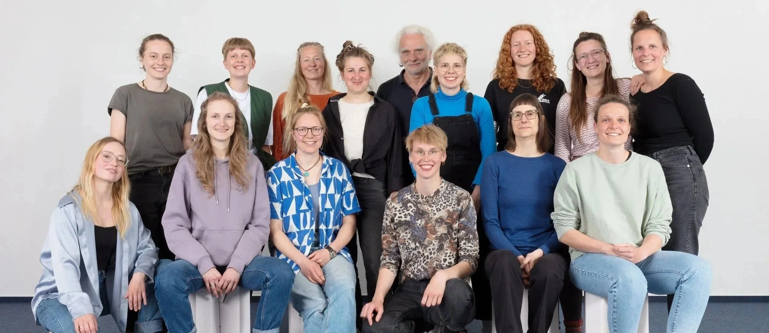 Auf dem Bild sieht man viele Menschen in einem hellen Raum. Sie stellen das Team der blau:pause dar. In erster Reihe sitzen Menschen und in der zweiten Reihe stehen Menschen.