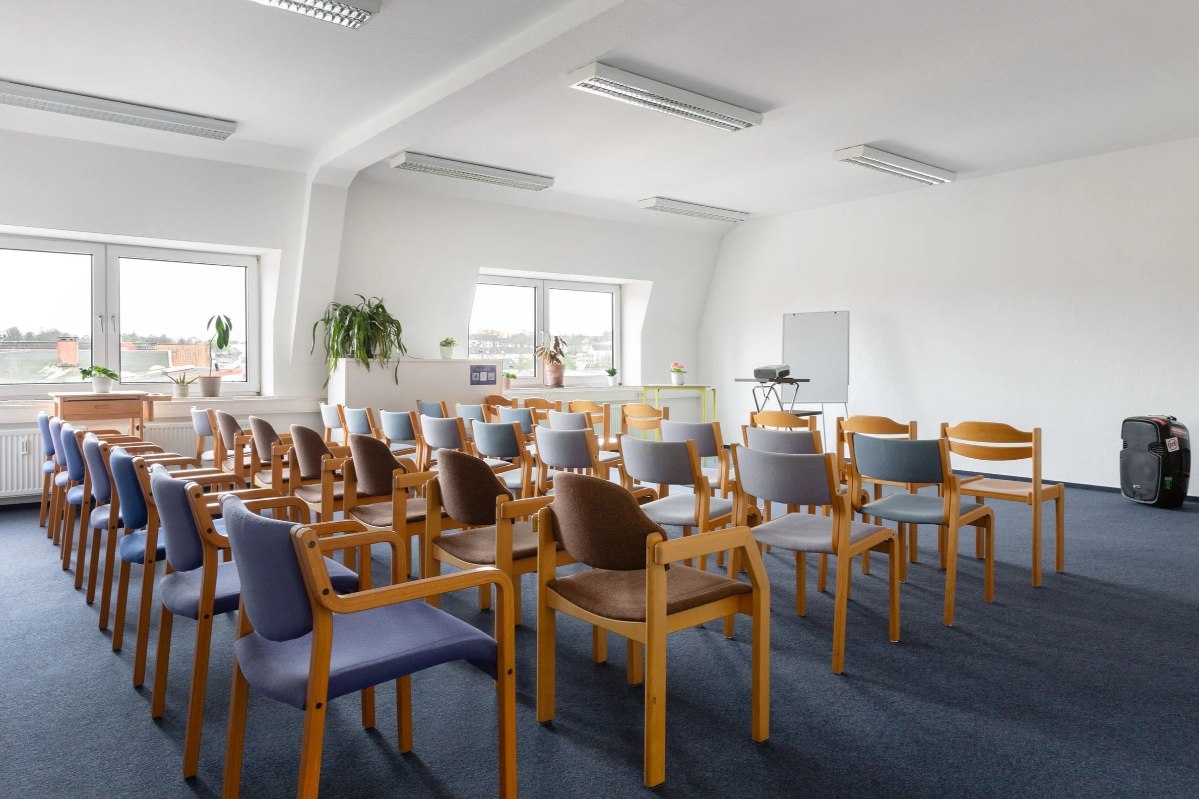 Das Bild zeigt einen großen Raum mit vielen Stühlen in einer Reihe. Die Wände sind weiß und die Stühle braun/blau.