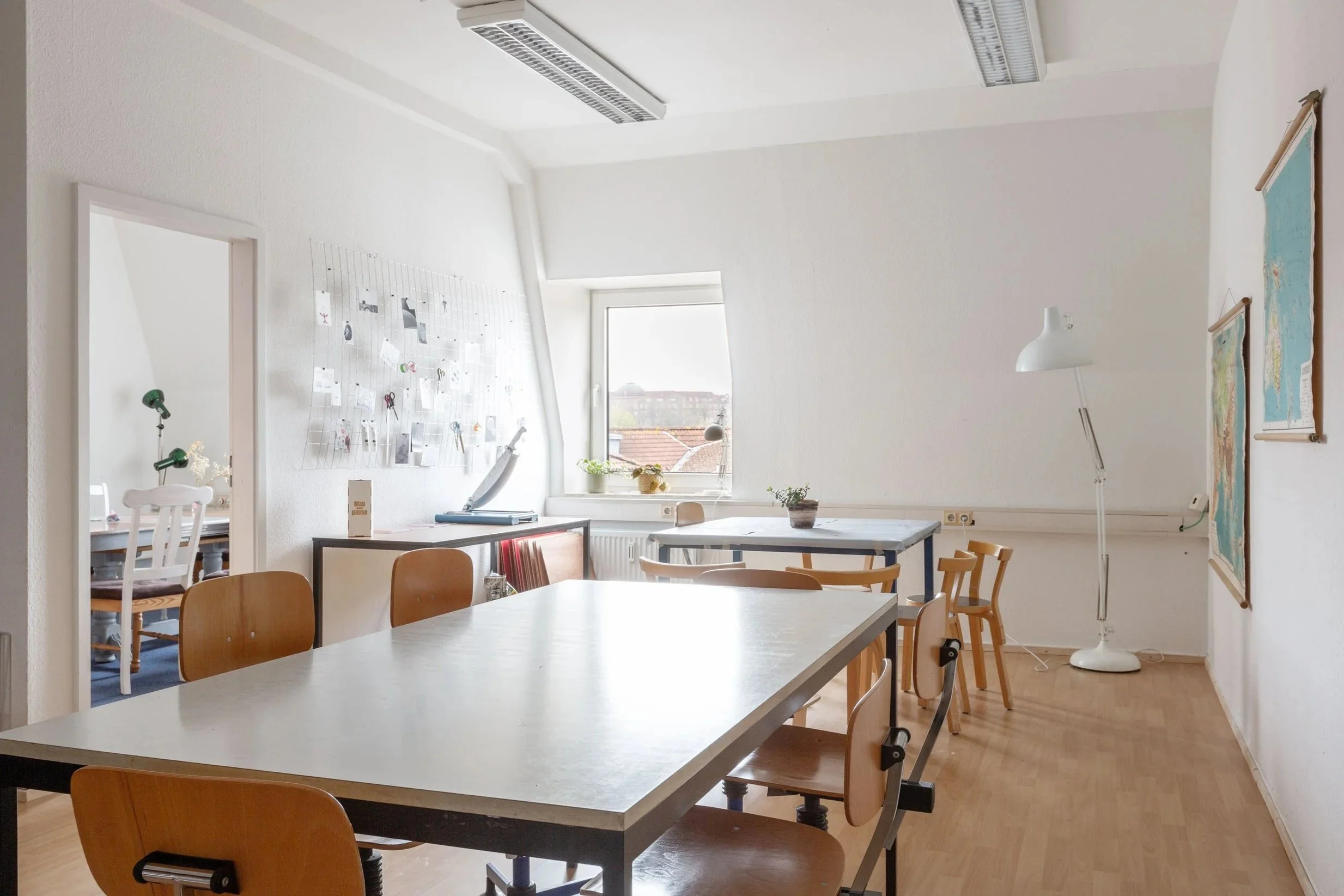 Auf dem Bild ist die kreativ:pause. Man sieht zwei weiße Tische mit vielen Stühlen Der Boden ist aus Holz und die Wände sind weiß.