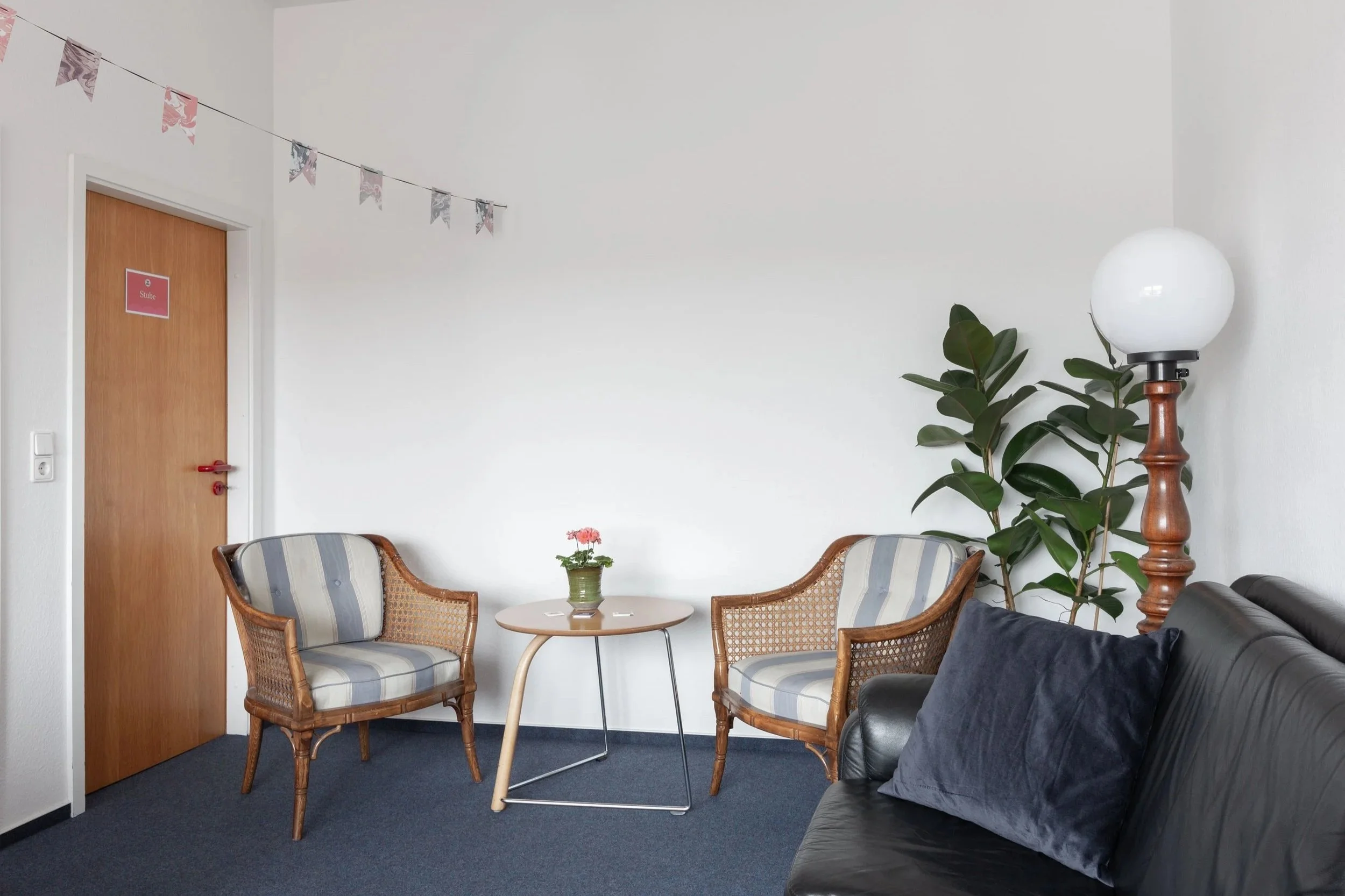 Auf dem Bild sieht man einen kleinen hellen Raum. Links ist eine braune Tür zu sehen und dann sieht man zwei Sessel und ein Sofa.