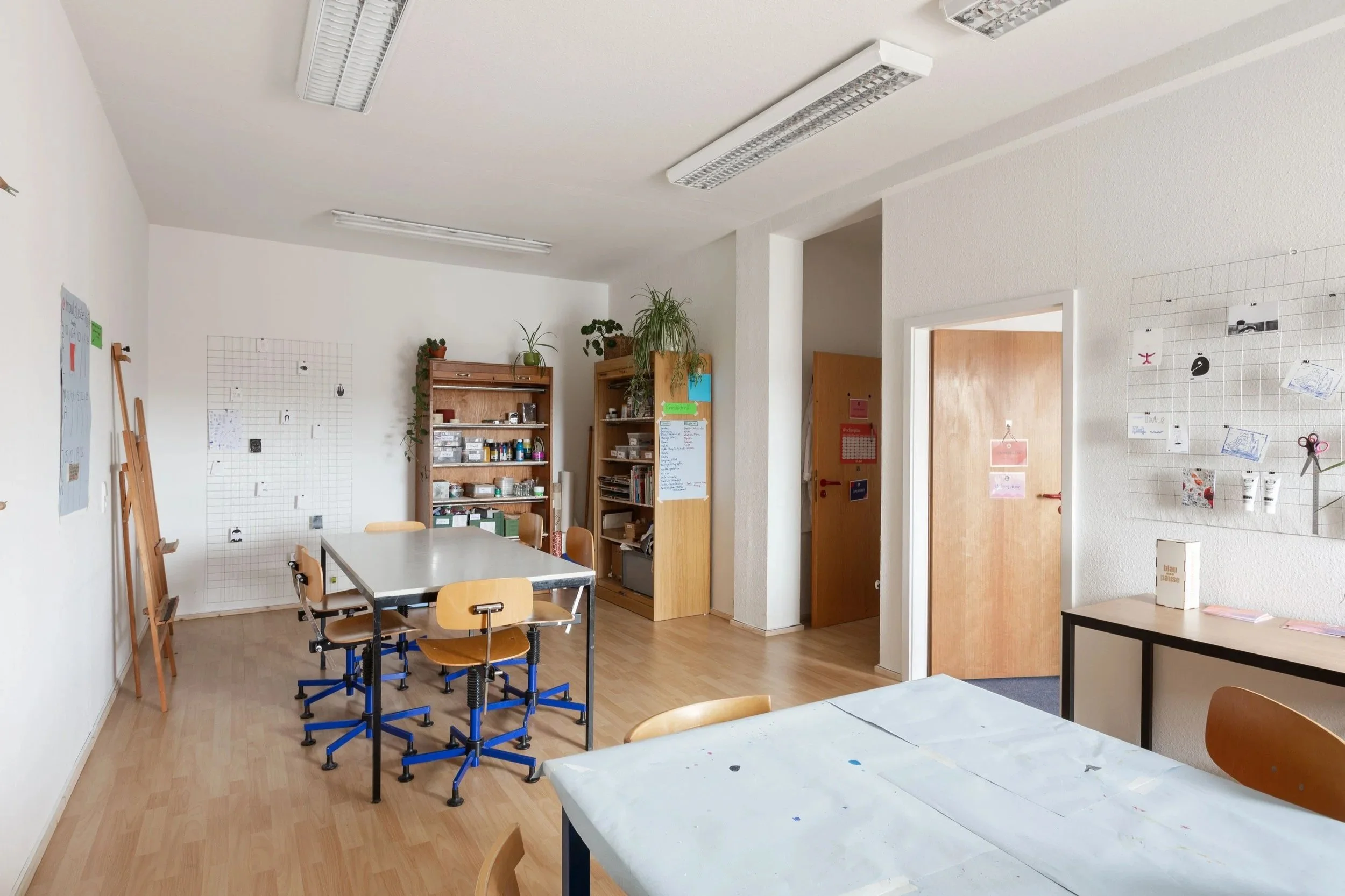 Auf dem Bild ist die kreativ:pause. Man sieht zwei weiße Tische mit vielen Stühlen Der Boden ist aus Holz und die Wände sind weiß. Im Hintergrund sieht man ein Regal mit Materialien.