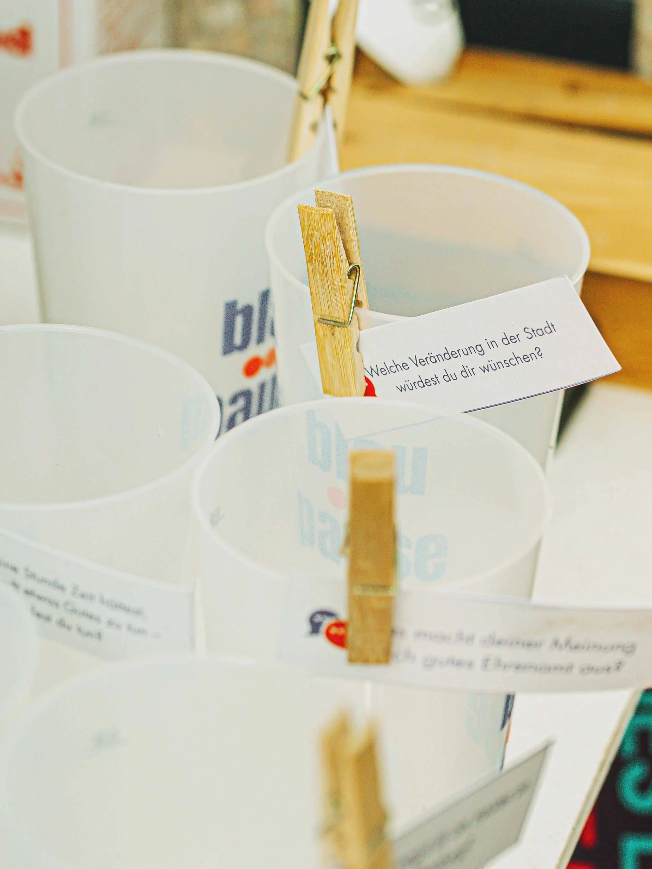 Auf dem Bild sind Becher zu sehen, an jedem Becher ist ein kleiner Zettel mit einer Wäscheklammer befestigt. Auf diesen Zetteln stehen Fragen.