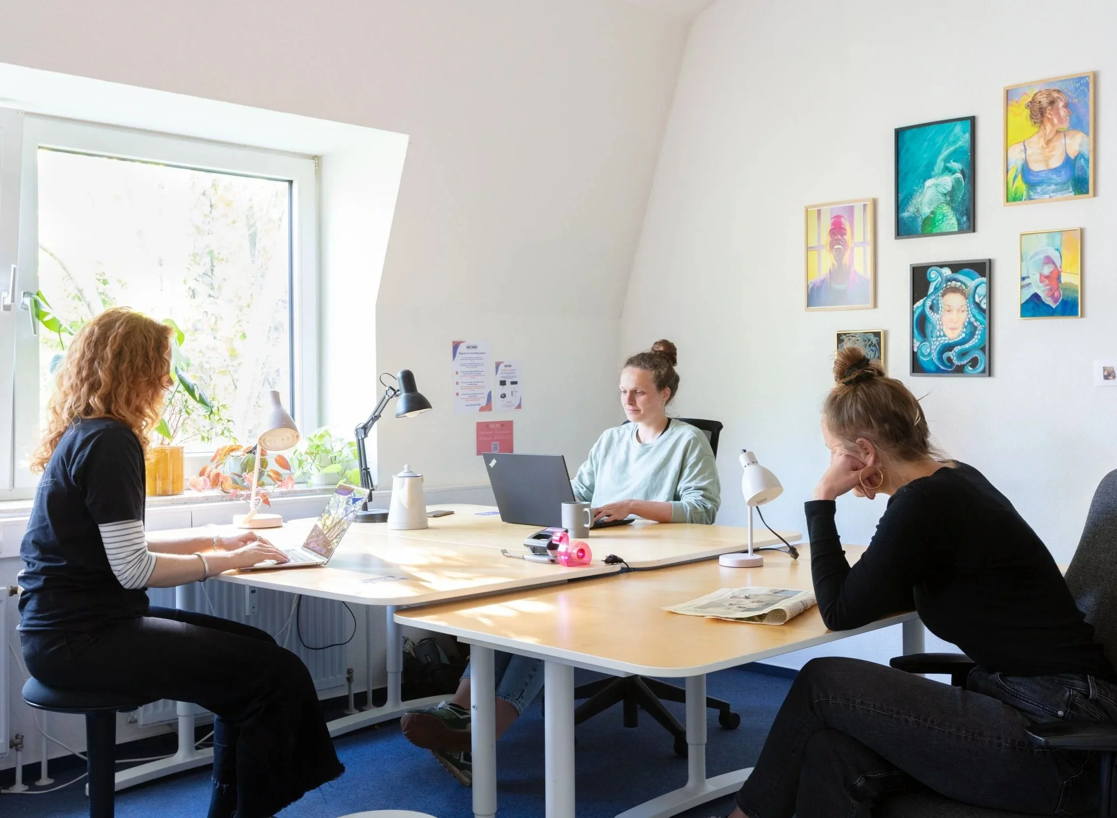 Das Bild zeigt drei Personen in einem Büro an einer Tischgruppe. Sie arbeiten.