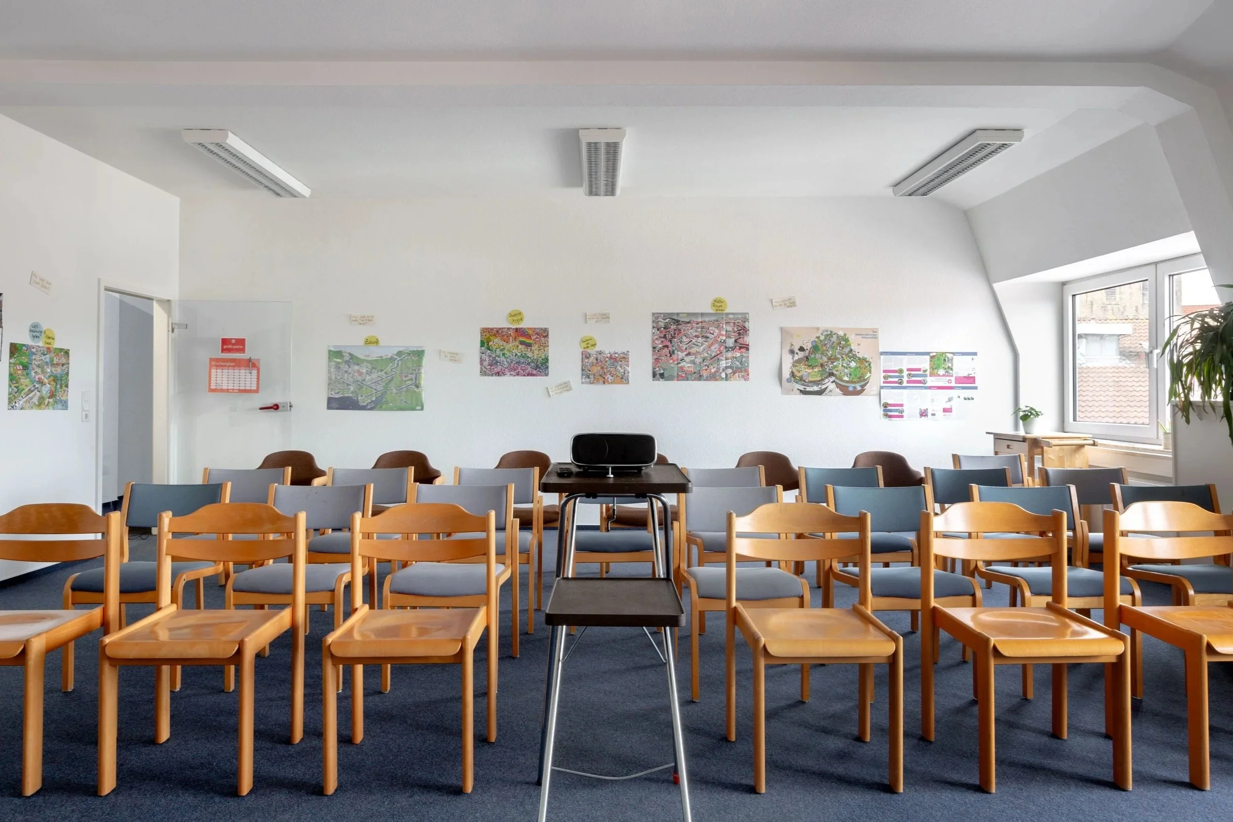 Das Bild zeigt einen großen Raum mit vielen Stühlen in einer Reihe. Die Wände sind weiß und die Stühle braun/blau.