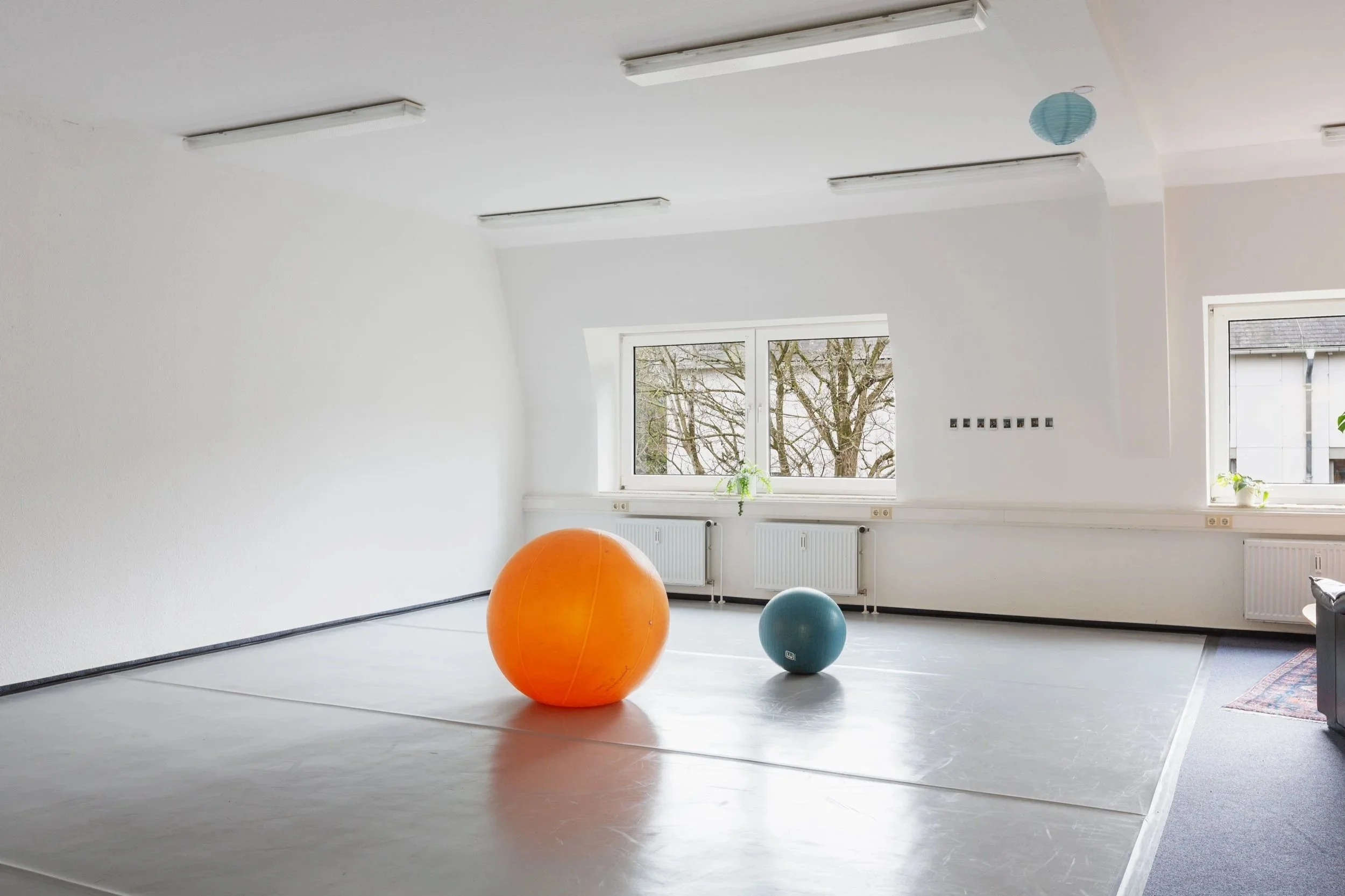 Hier sieht man die bewegungs:pause. Ein großer heller Raum mit einem grauen Tanzboden. Auf der Tanzfläche sind zwei große Bälle zu sehen.