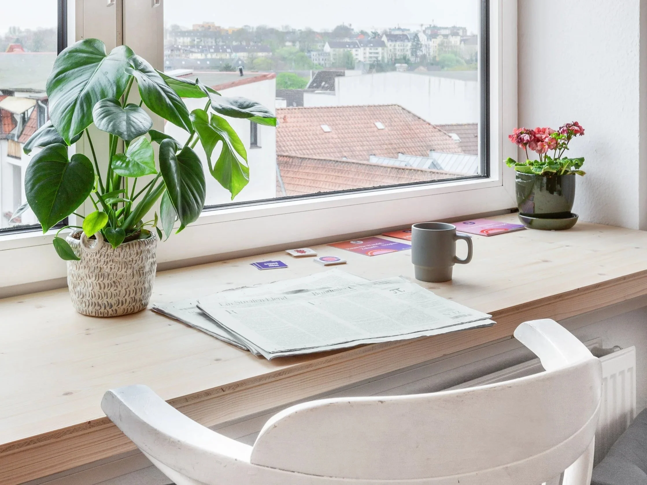Auf de Bild sieht man eine Holzfensterbank. Auf dieser liegt eine Zeitung und Sticker und es steht eine Kaffetasse daneben. Man kann nach draußen schauen und sieht Flensburg.