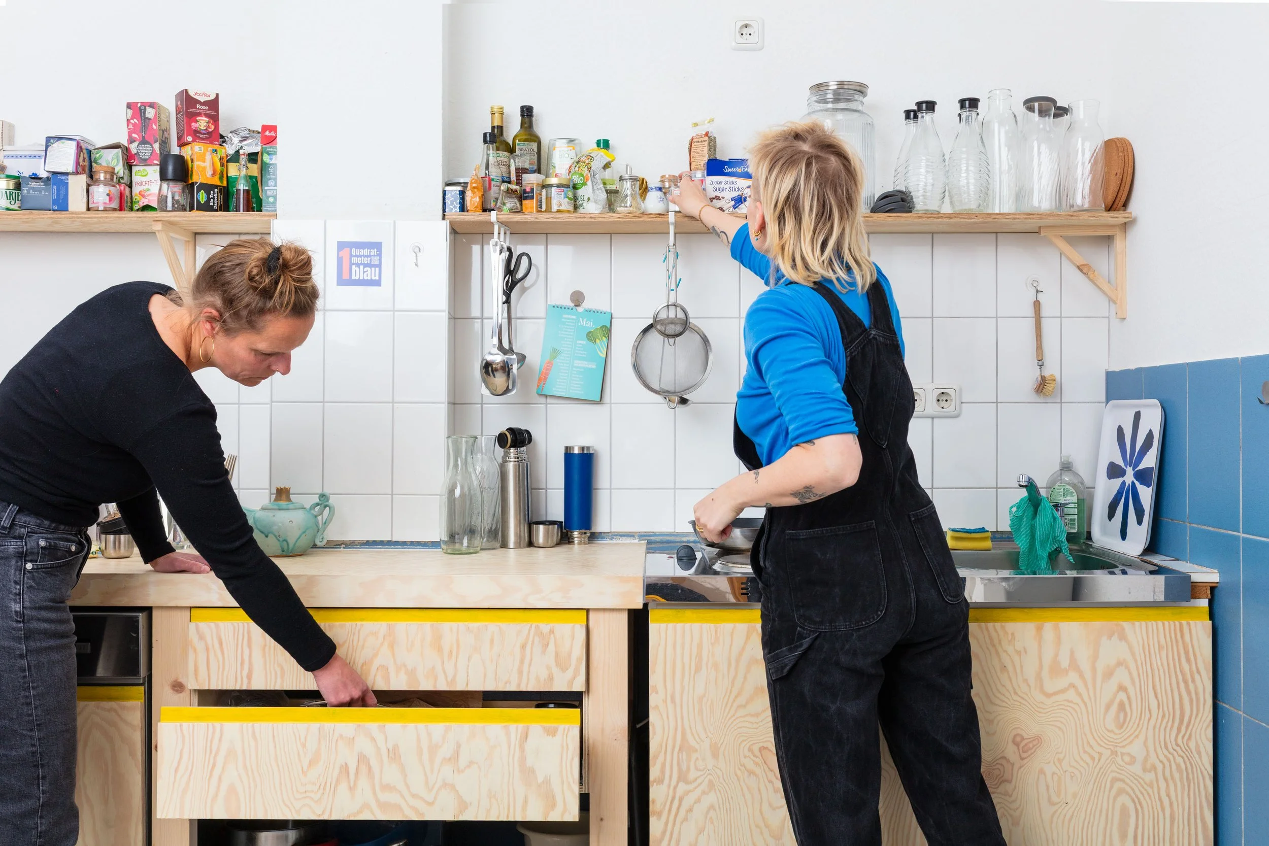 Auf dem Bild sieht man eine helle Holzküche und zwei Personen von hinten, die diese einräumen.