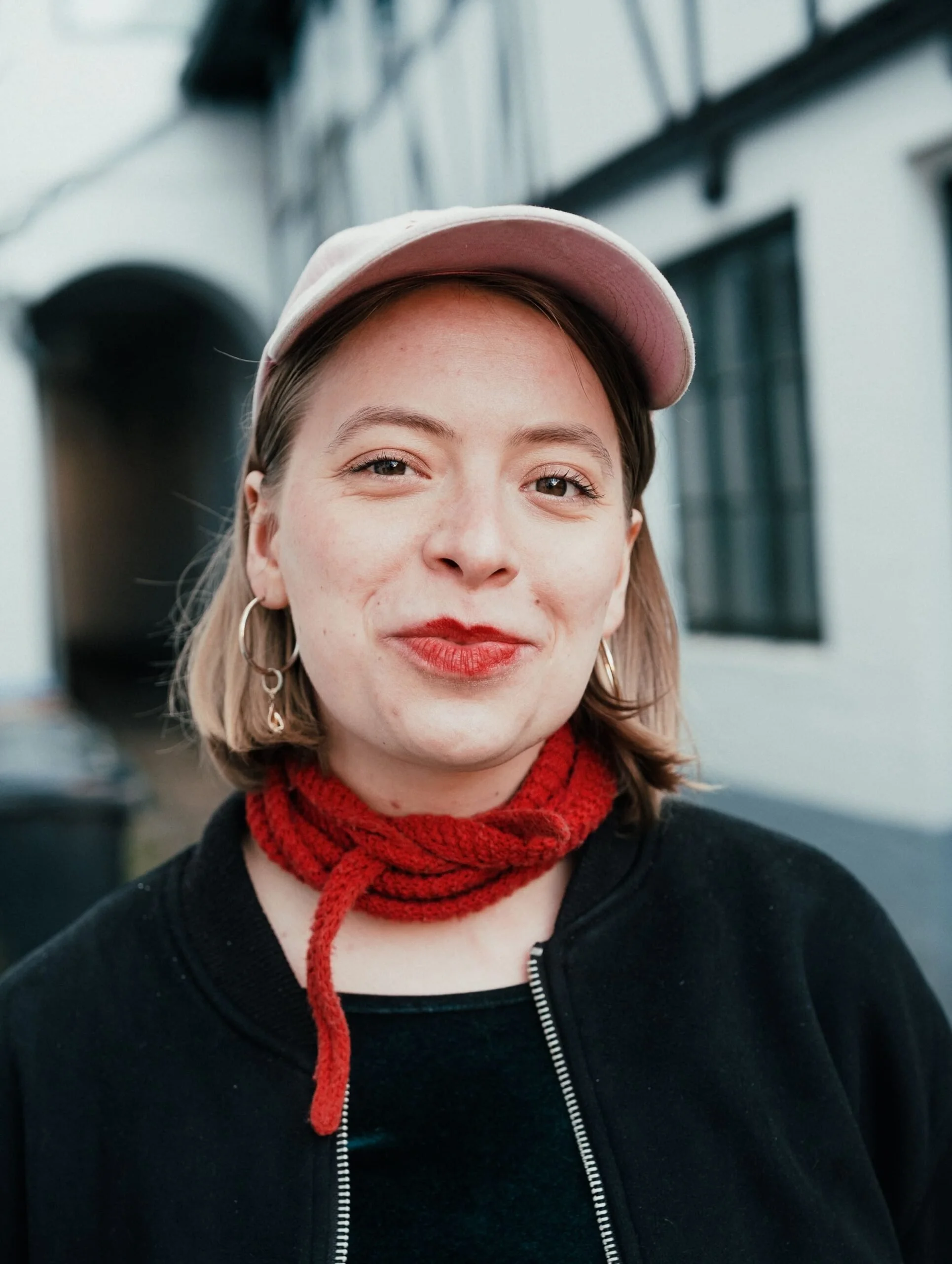 Nahaufnahme eines lachenden jungen Mädchens mit roten Lippen, Ohrringen, einer rosa Basecap und einer roten Strickschleife um den Hals, davor ein dunkler Mantel, im Hintergrund ein weißes Gebäude mit schwarzen Fenstern.