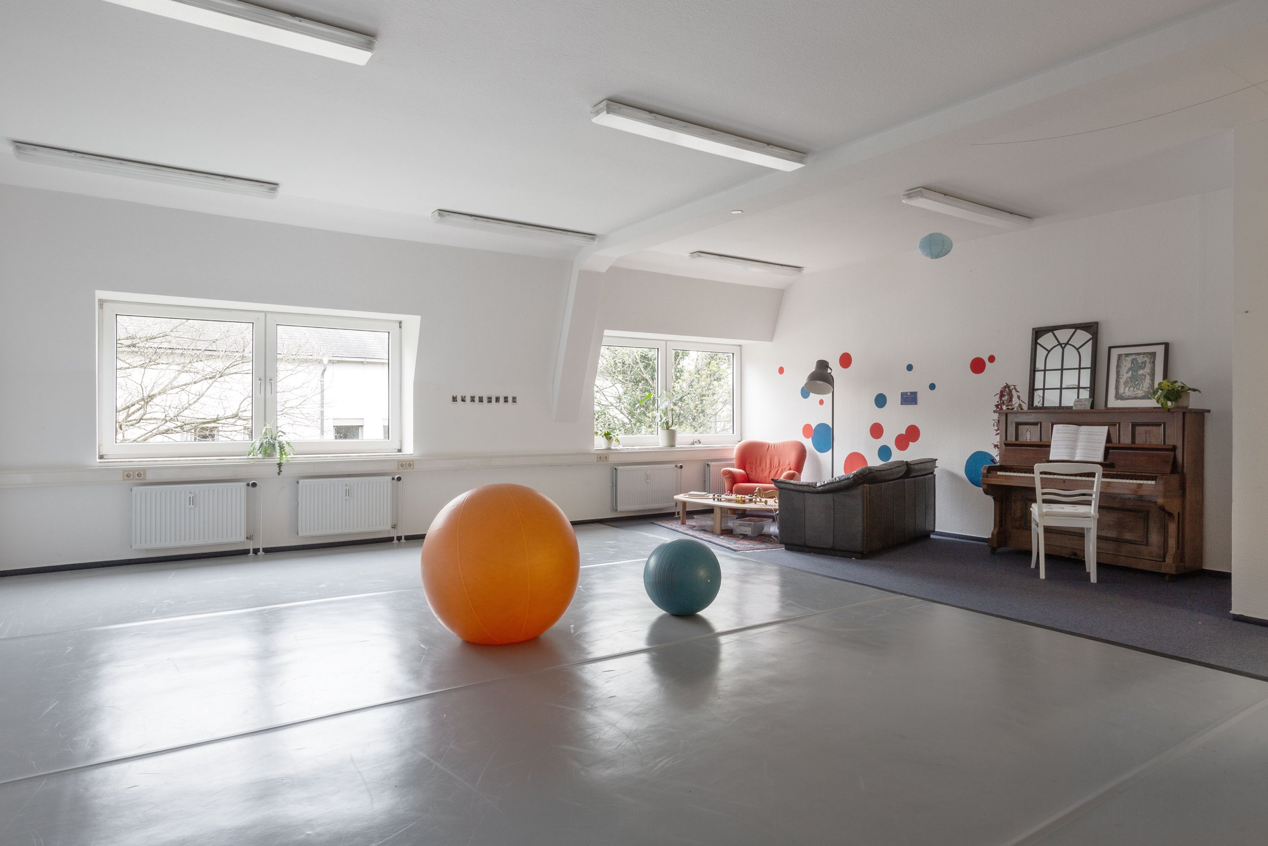 Hier sieht man die bewegungs:pause. Ein großer heller Raum mit einem grauen Tanzboden. Im Hintergrund sieht man ein Klaiver und eine Sofaecke. Auf der Tanzfläche sind zwei große Bälle zu sehen.