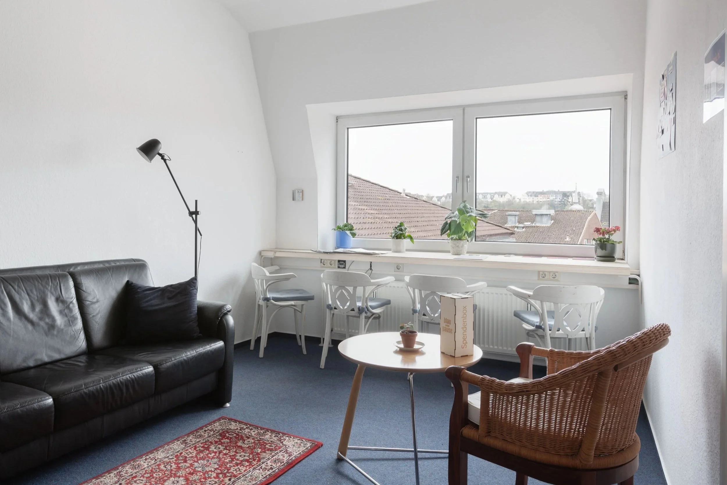 Es ist ein kleiner Raum zu siehen. Links steht ein schwarzes Sofa. Im Hintergrund sieht man eine Holzfensterbank mit vier Stühlen und ein Blick über die Dächer von Flensburg.