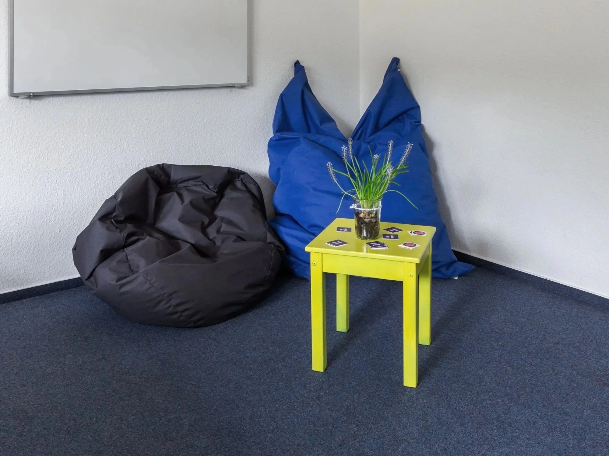 Auf dem Bild sieht man einen blauen und einen schwarzen Sitzsack in einem Raum. Davor steht ein gelber Tisch mit einer Blume drauf.