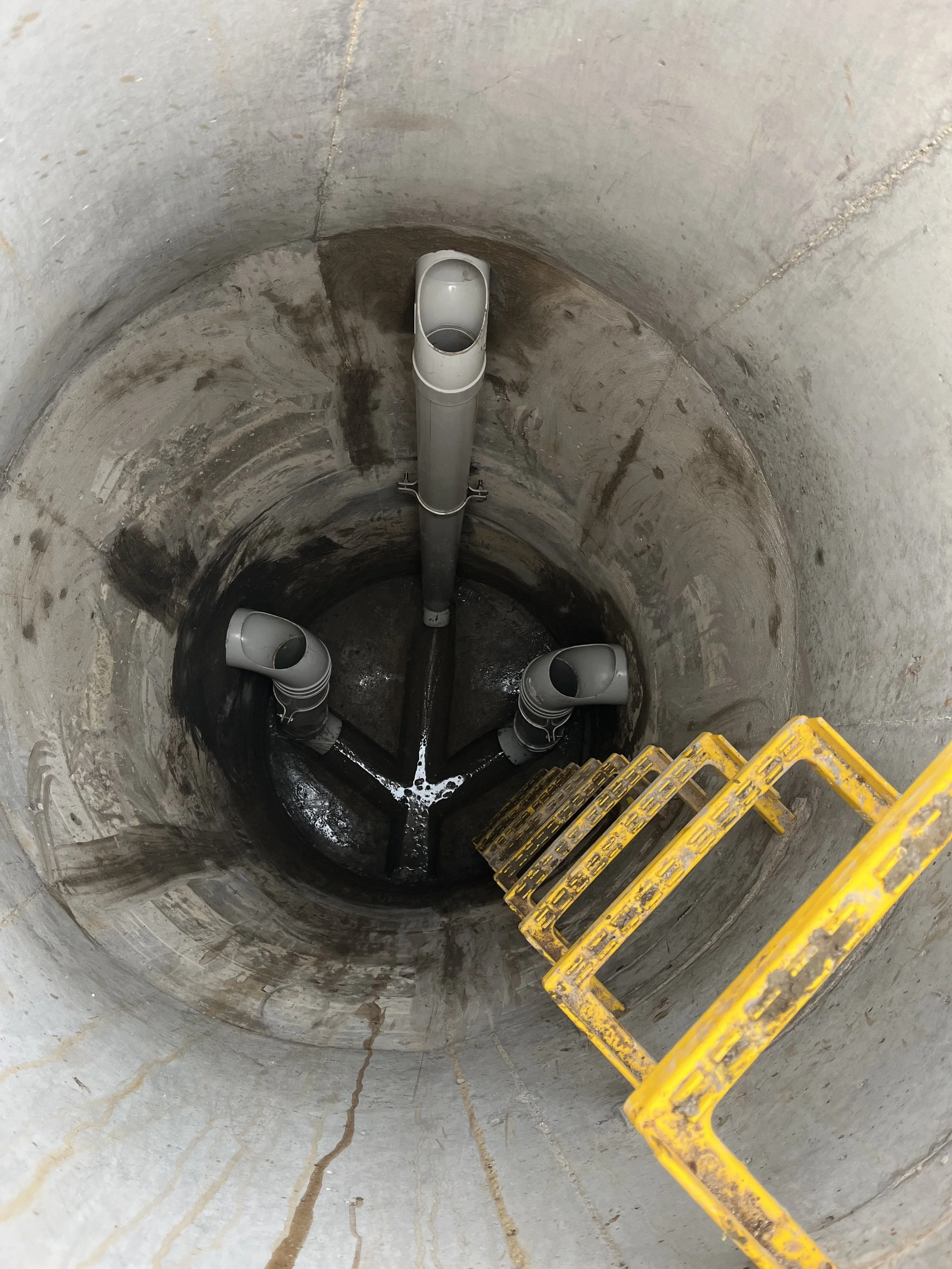 View down a sewer or utility shaft with three pipes at the bottom, concrete walls, and a yellow ladder descending into the shaft.