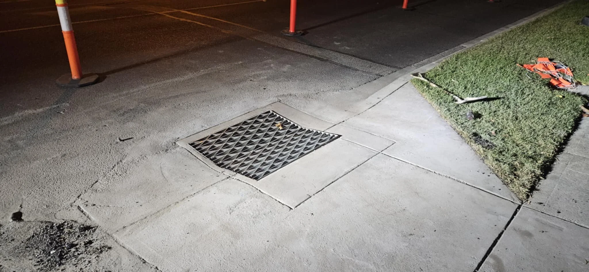 Street sidewalk with a storm drain, construction cones, and a small patch of grass with a fallen orange construction vest.