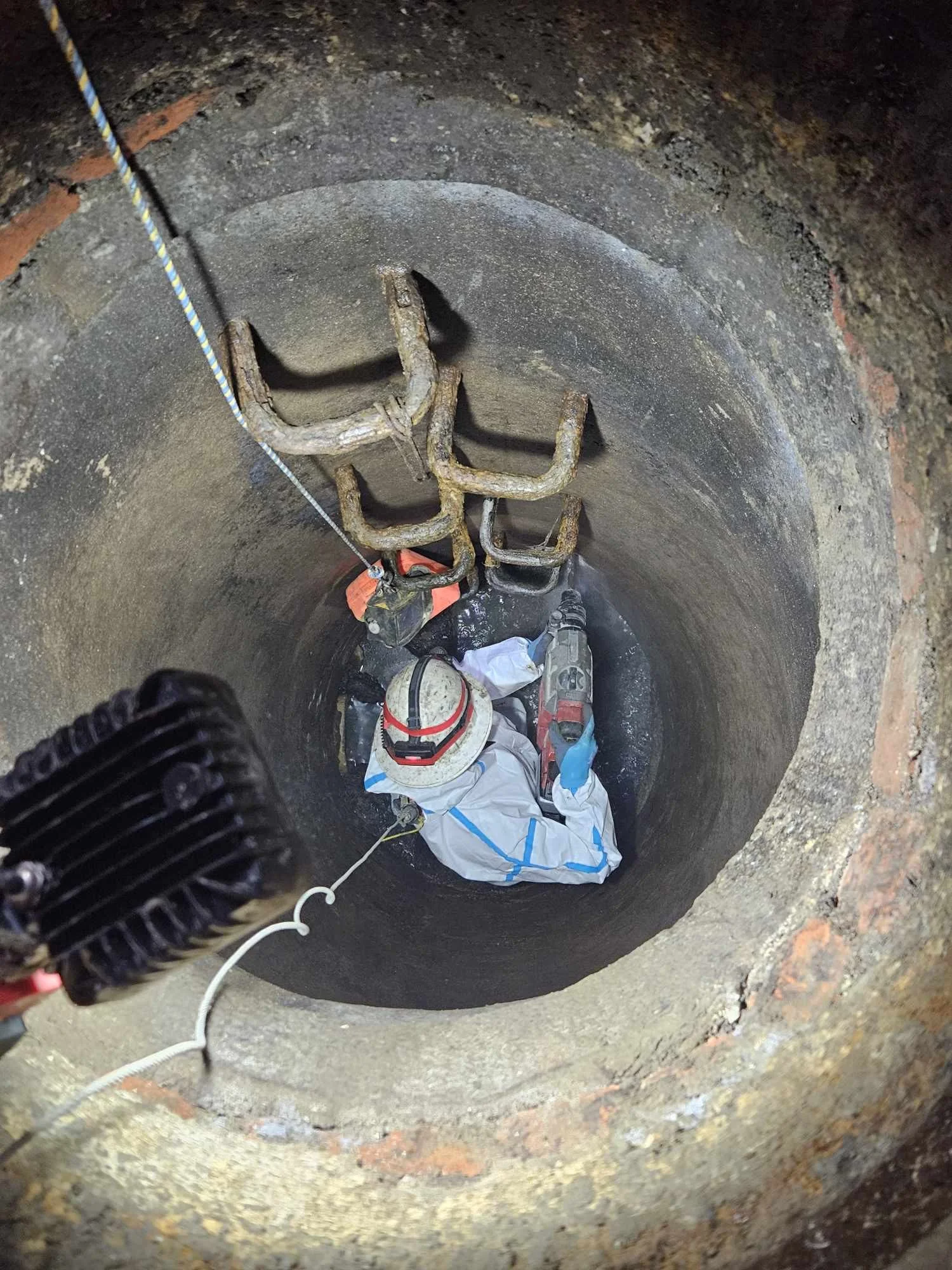 Worker in protective gear inside a deep construction or maintenance pit with metal rungs and a rope ladder.