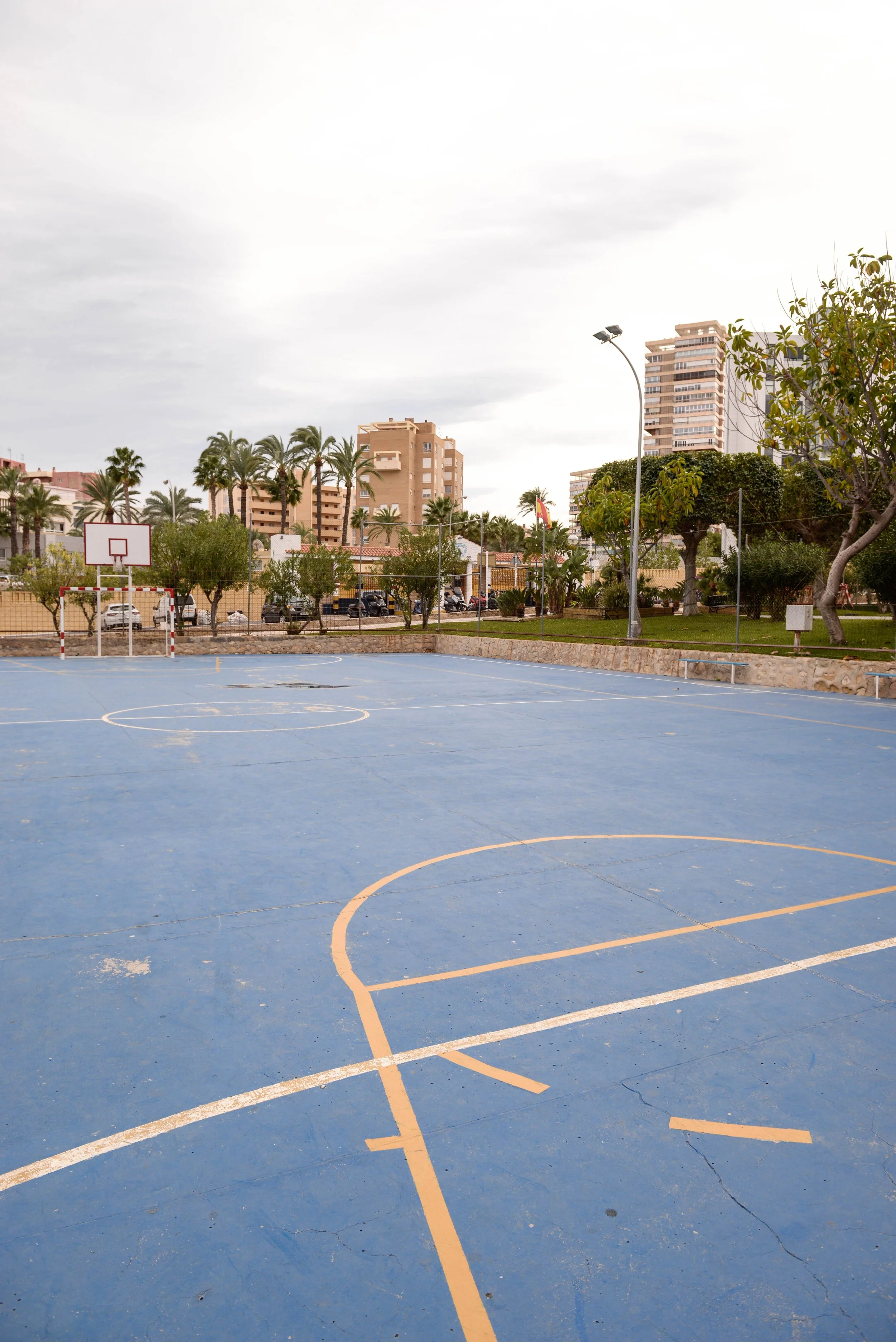 Terrain de basketball extérieur avec des marques jaunes sur un sol bleu, entouré d'arbres, de bâtiments résidentiels et d'une clôture, sous un ciel nuageux.