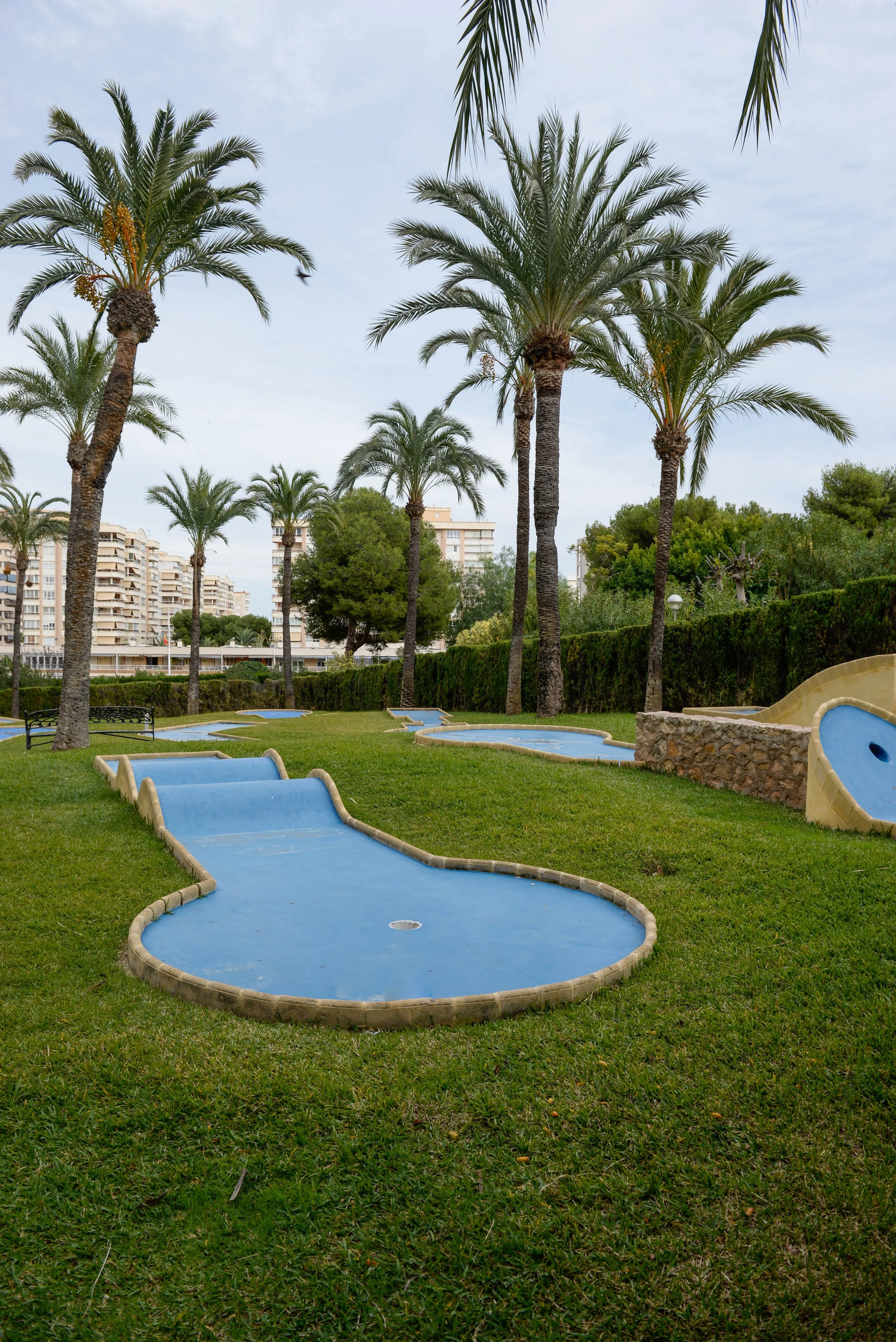 Terrain de mini-golf avec plusieurs trous et obstacles, entouré de palmiers et de pelouse verte, avec des bâtiments en arrière-plan.