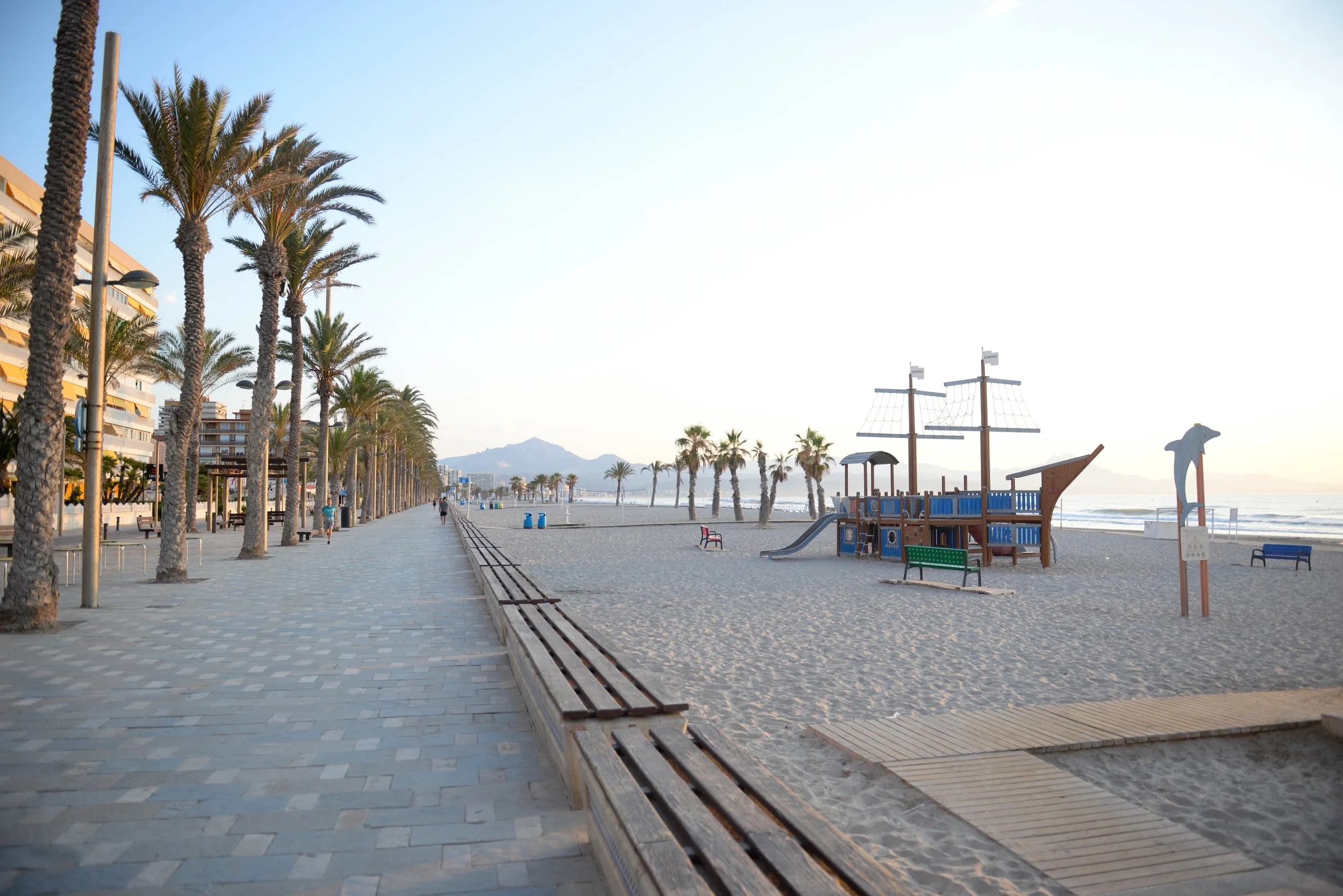 Promenade piétonne en bord de mer avec palmiers, plage de sable, installations pour enfants en forme de bateau pirate et chiens, avec montagnes au loin, en fin d'après-midi.