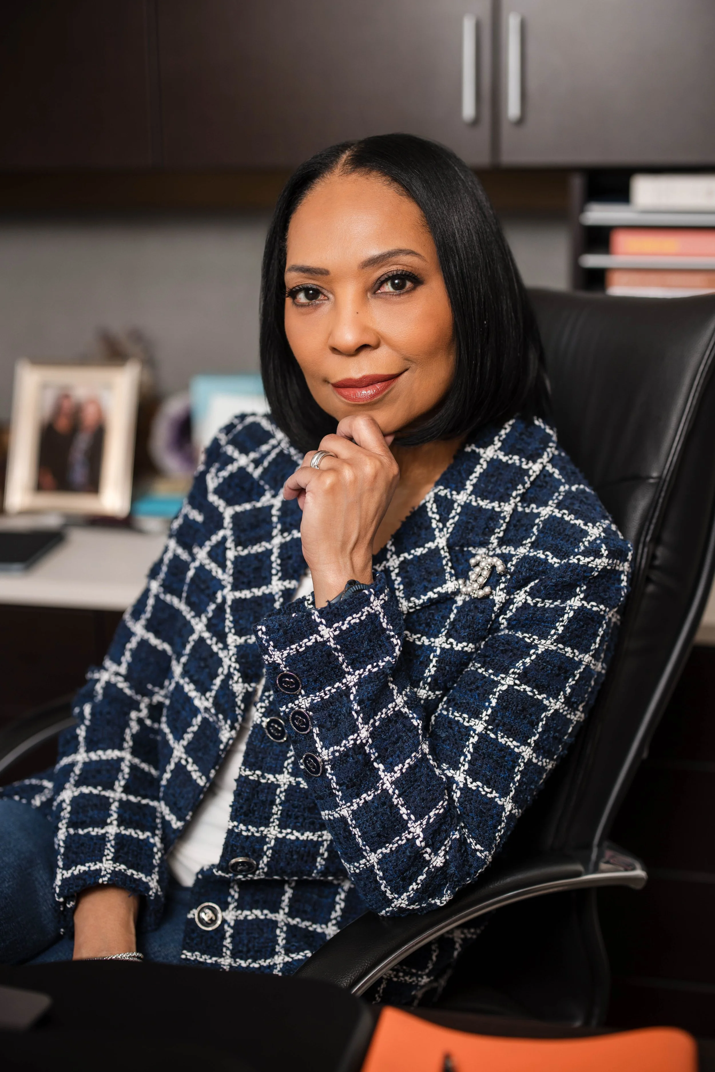 A woman sitting in an office chair with her hand near her chin, smiling softly. She has shoulder-length black hair, wears a blue and white checkered blazer, and is in an office setting with shelves, framed photographs, and organized files in the background.